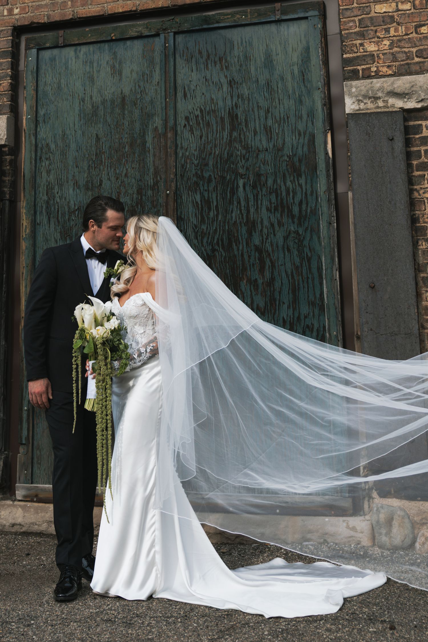 Bride and groom kiss outdoors with flowing cathedral veil billowing beside rustic green doors.