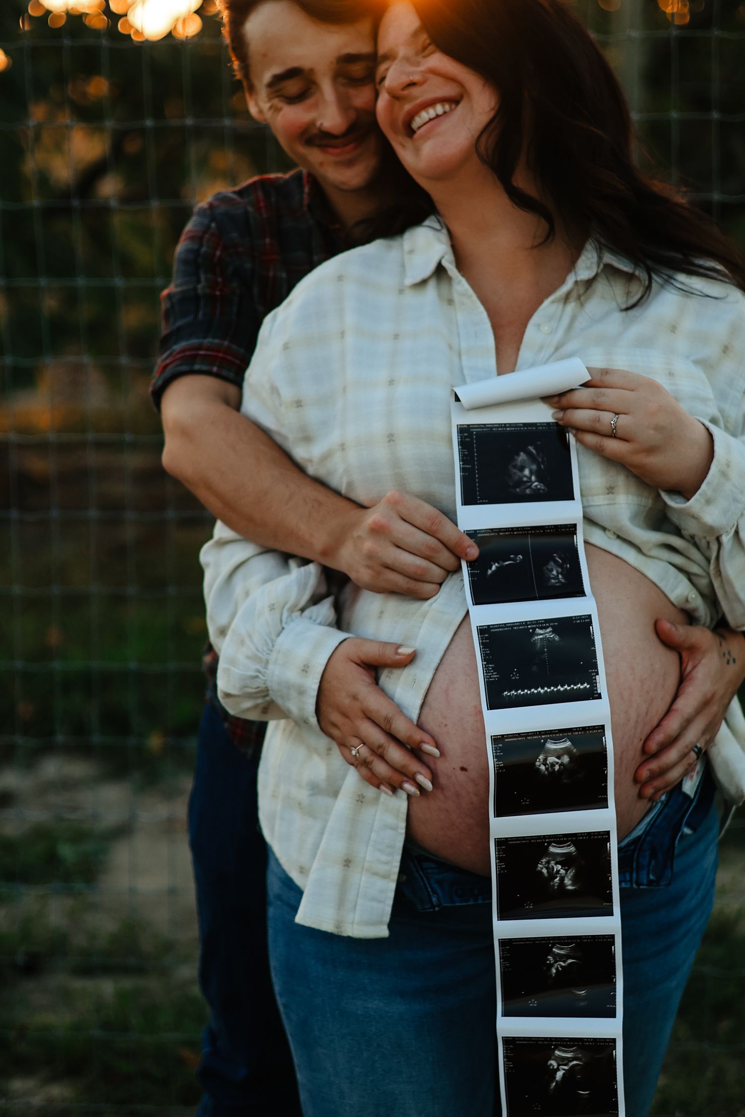 Pregnant couple holding ultrasound photos at golden hour, woman smiling joyfully while partner embraces her from behind.