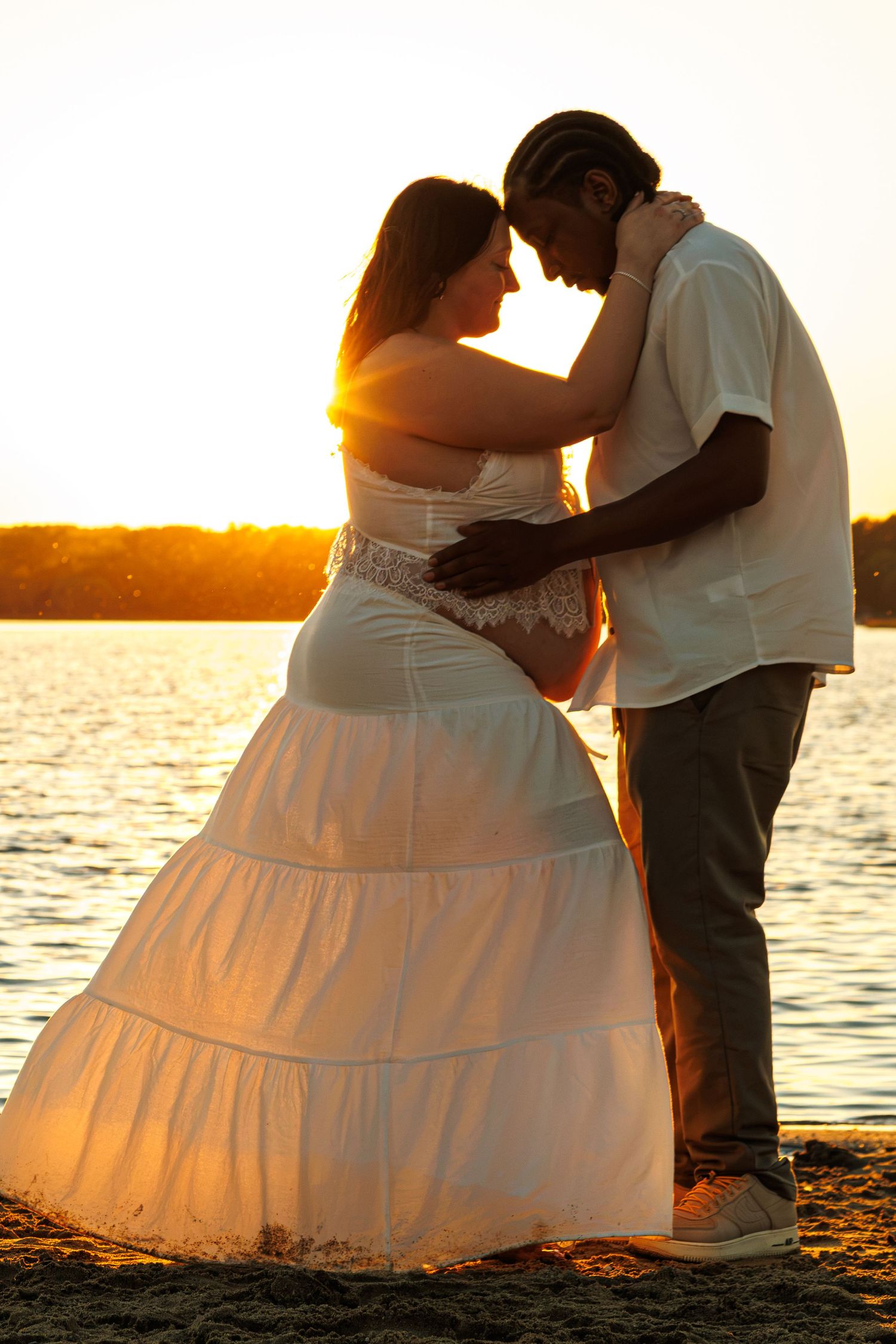 Interracial couple embracing tenderly at golden sunset by the lake, pregnant woman in white dress.