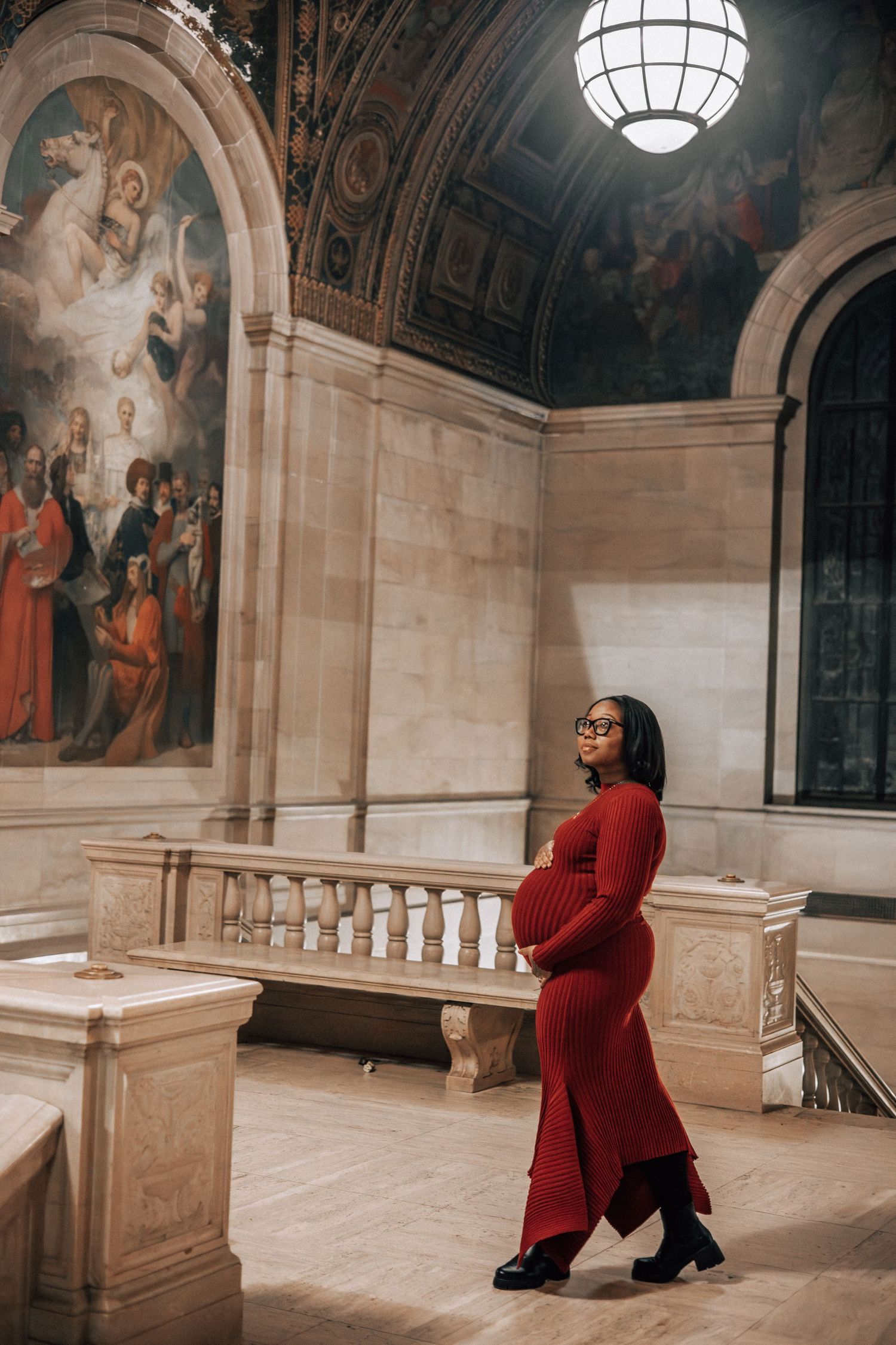 Pregnant woman in red maternity dress posing under chandelier in stunning classical architecture hall.