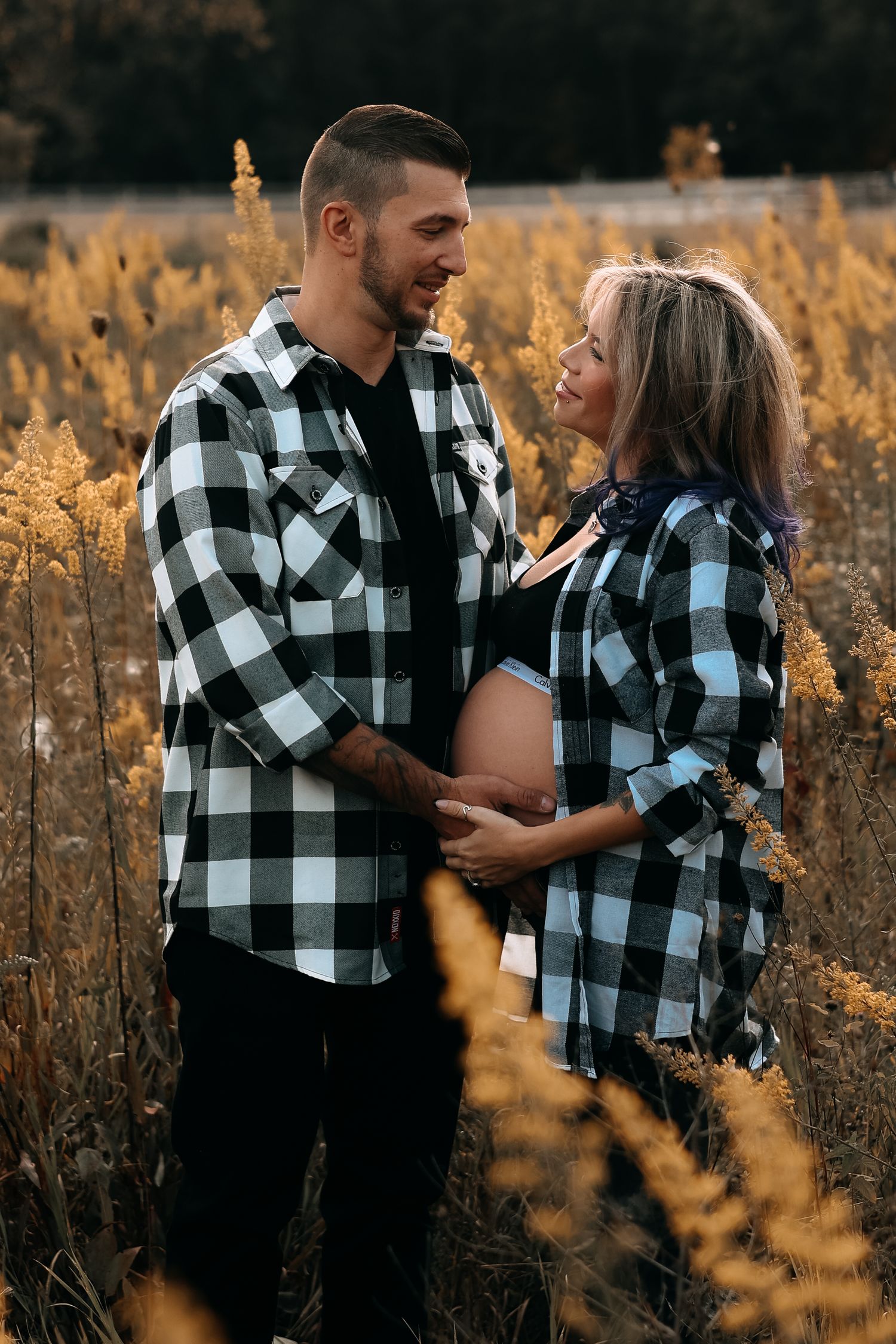 Couple in matching plaid shirts embracing in autumn field, woman showing pregnant belly outdoors.