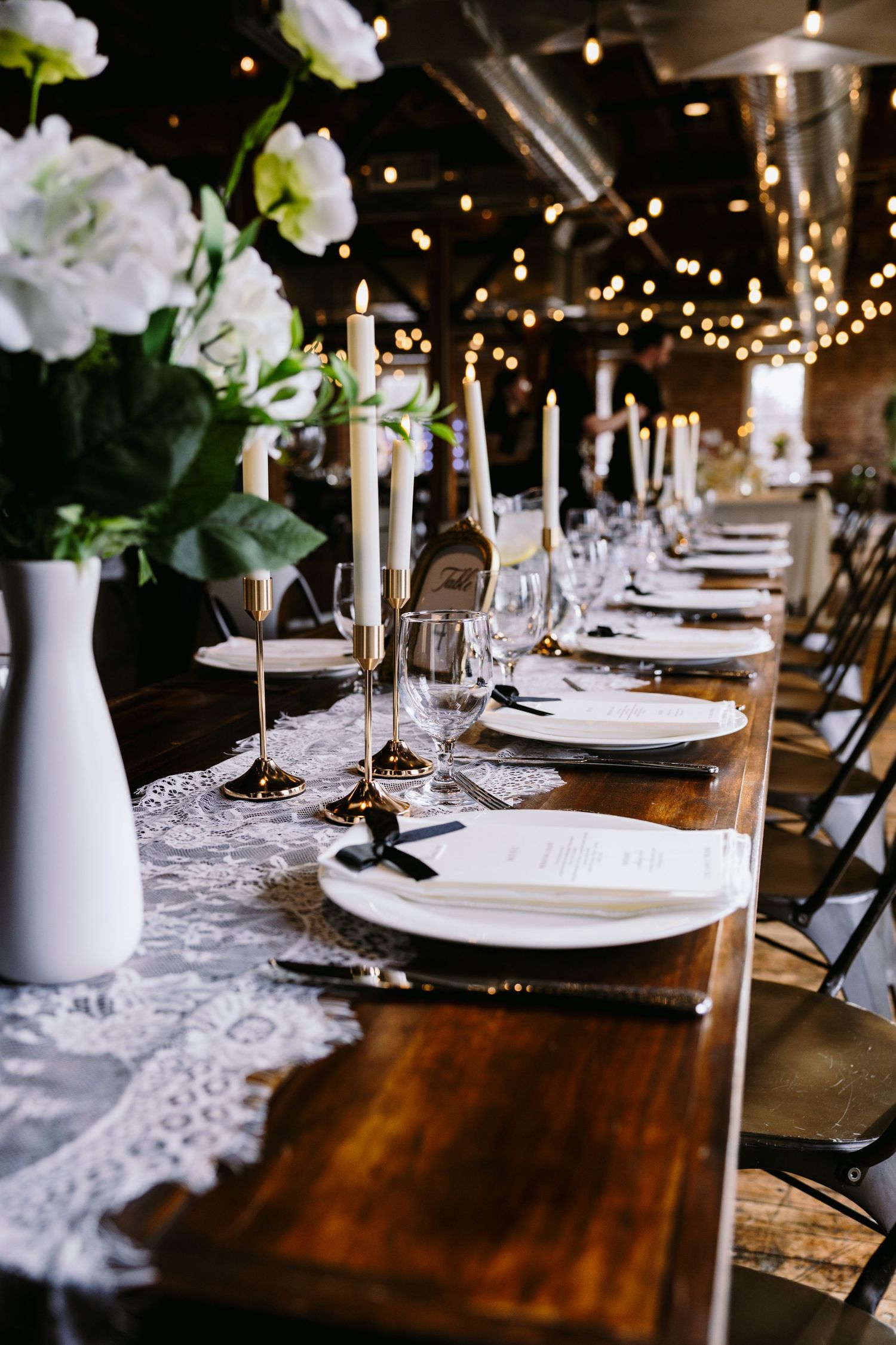 Rustic wedding reception table with white florals, gold candlesticks, lace runner, and string lights.