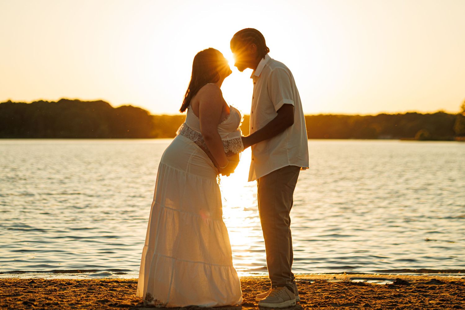 Pregnant couple silhouetted against golden sunset by a lake, sharing a tender moment in white outfits.
