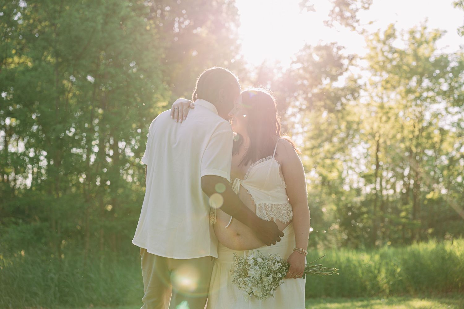 Couple in white embracing outdoors with golden sunlight filtering through lush green trees, holding a bridal bouquet.