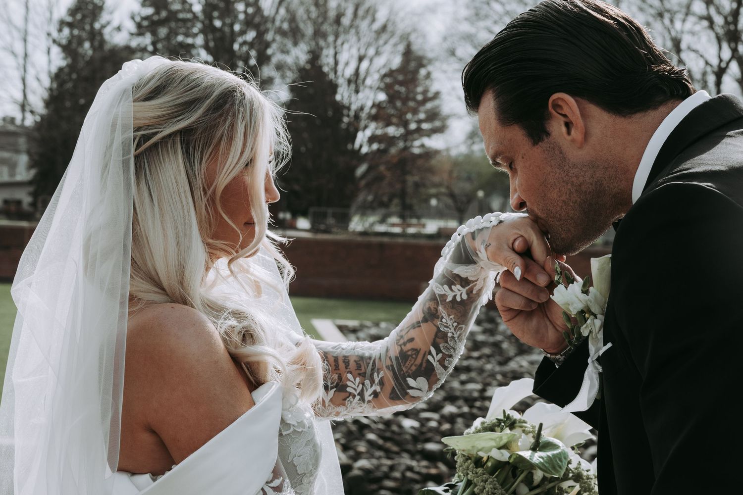 Groom tenderly kisses brides lace-gloved hand during romantic outdoor wedding portrait session.