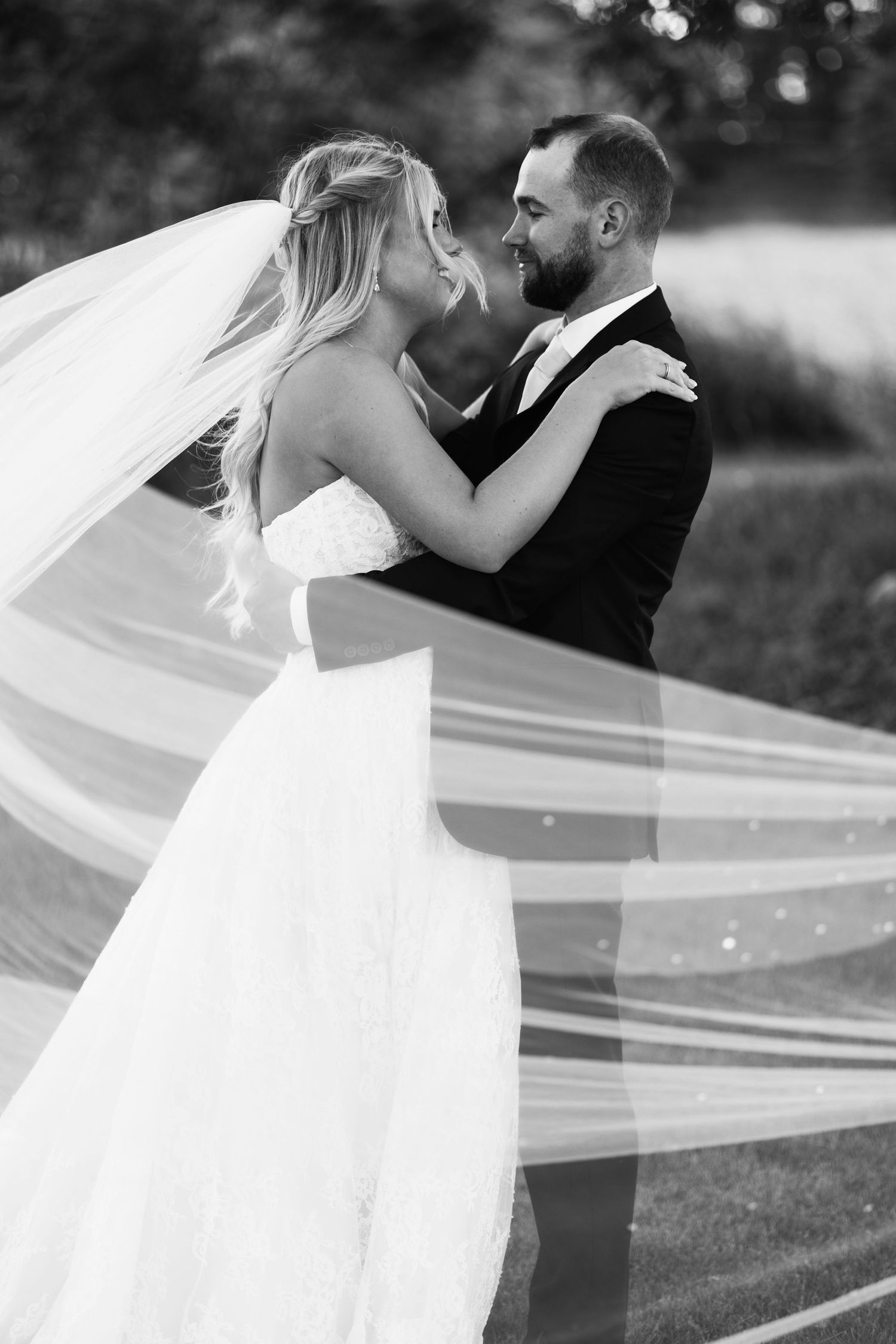 Black and white photo of bride and groom embracing, veil flowing in wind.
