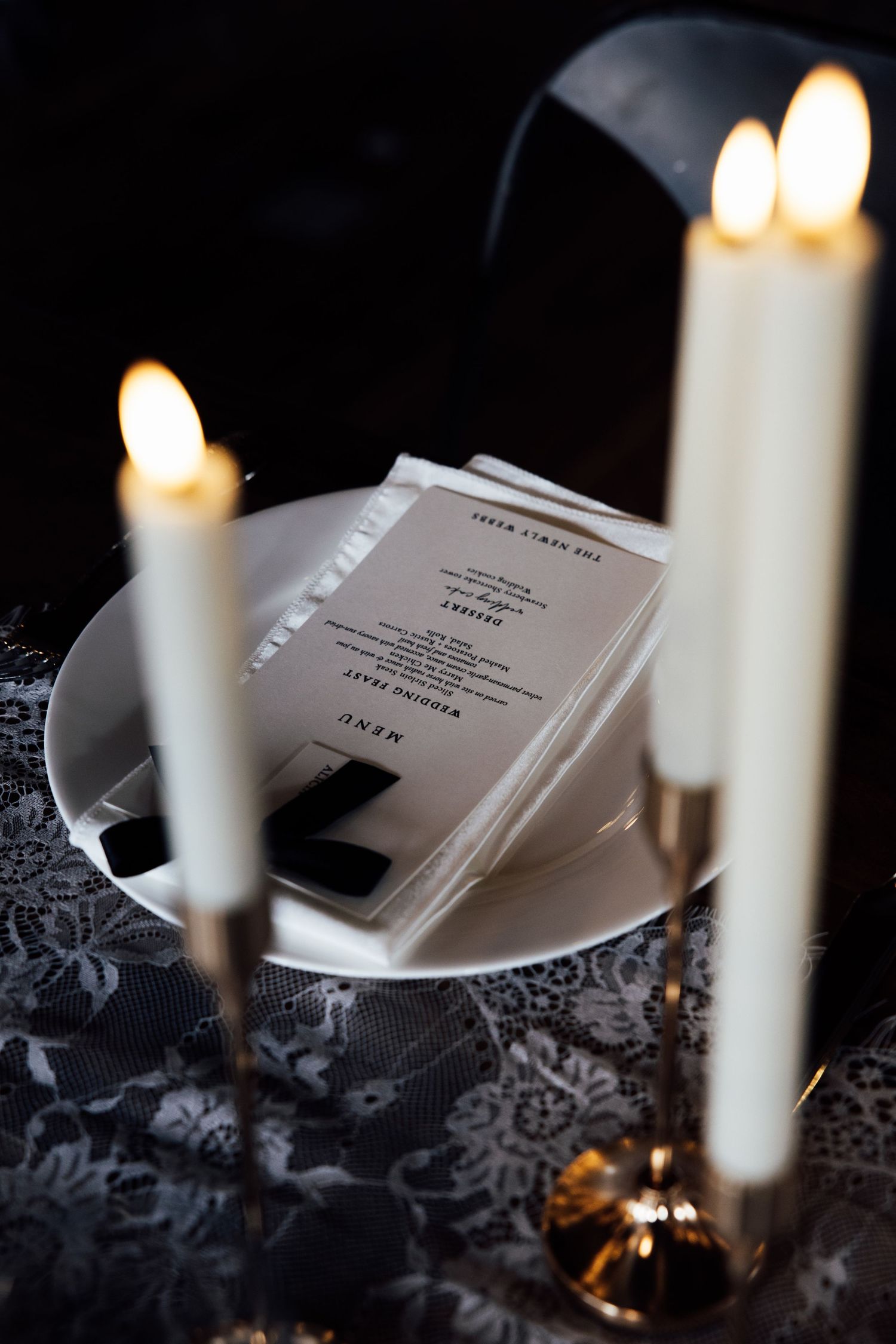 Elegant wedding menu cards on white plate surrounded by tall white candles on lace tablecloth.
