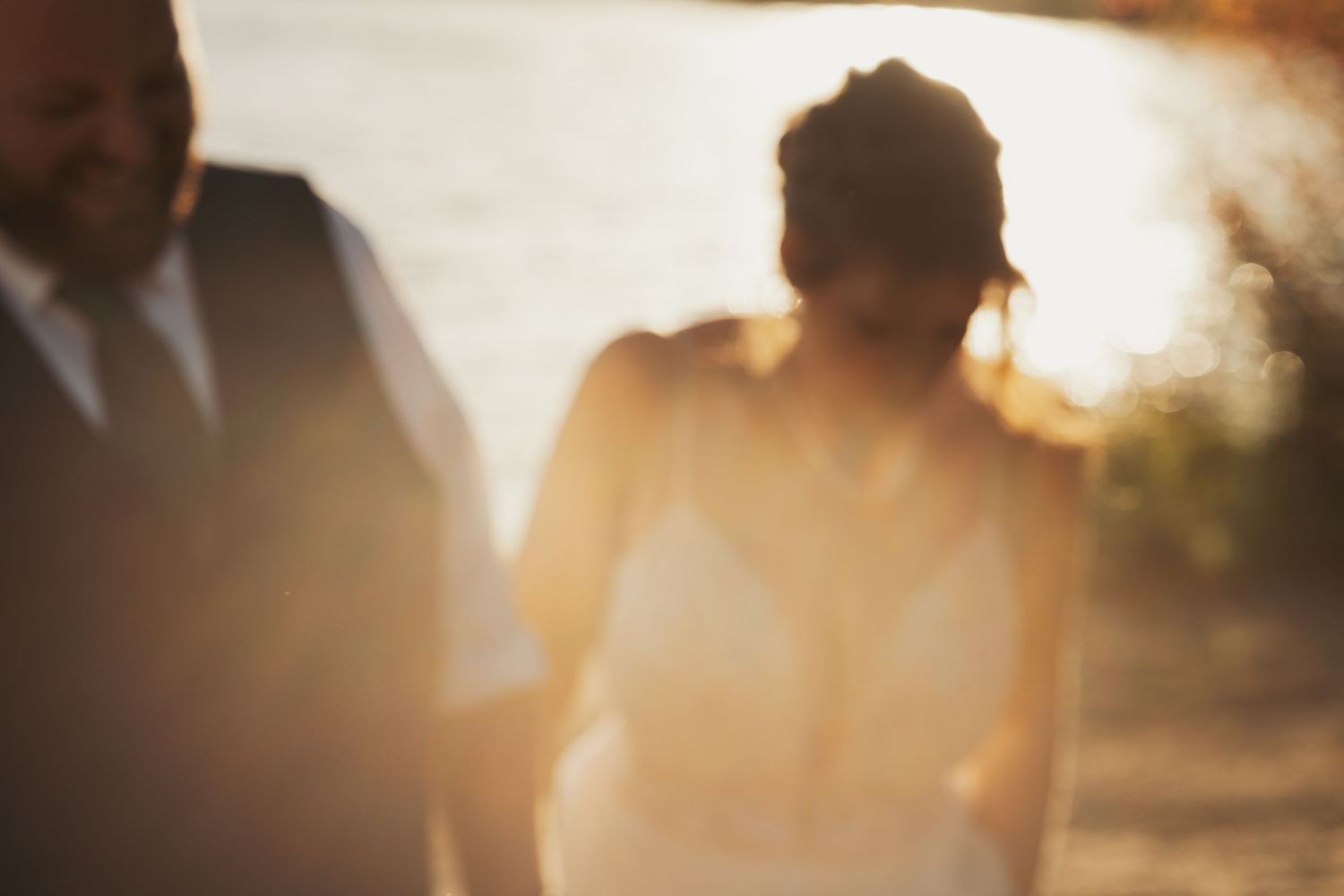 A couple in wedding attire walks hand in hand at golden hour near a sunlit lake.