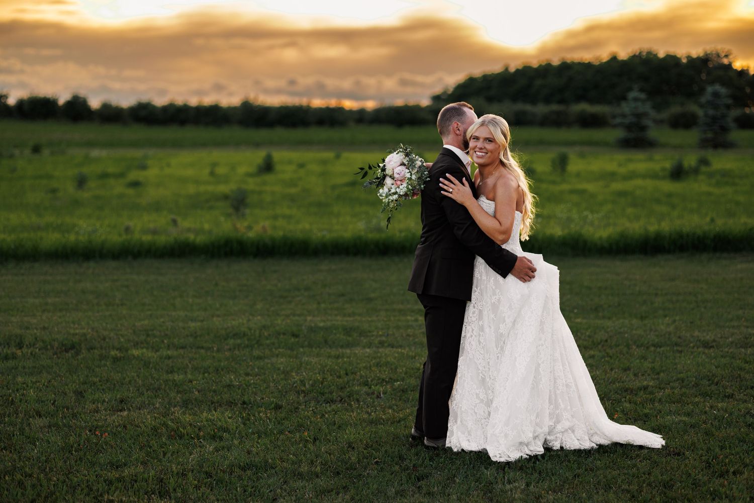 Bride and groom embrace in lush green field at golden sunset, bride holding white floral bouquet in wedding dress.