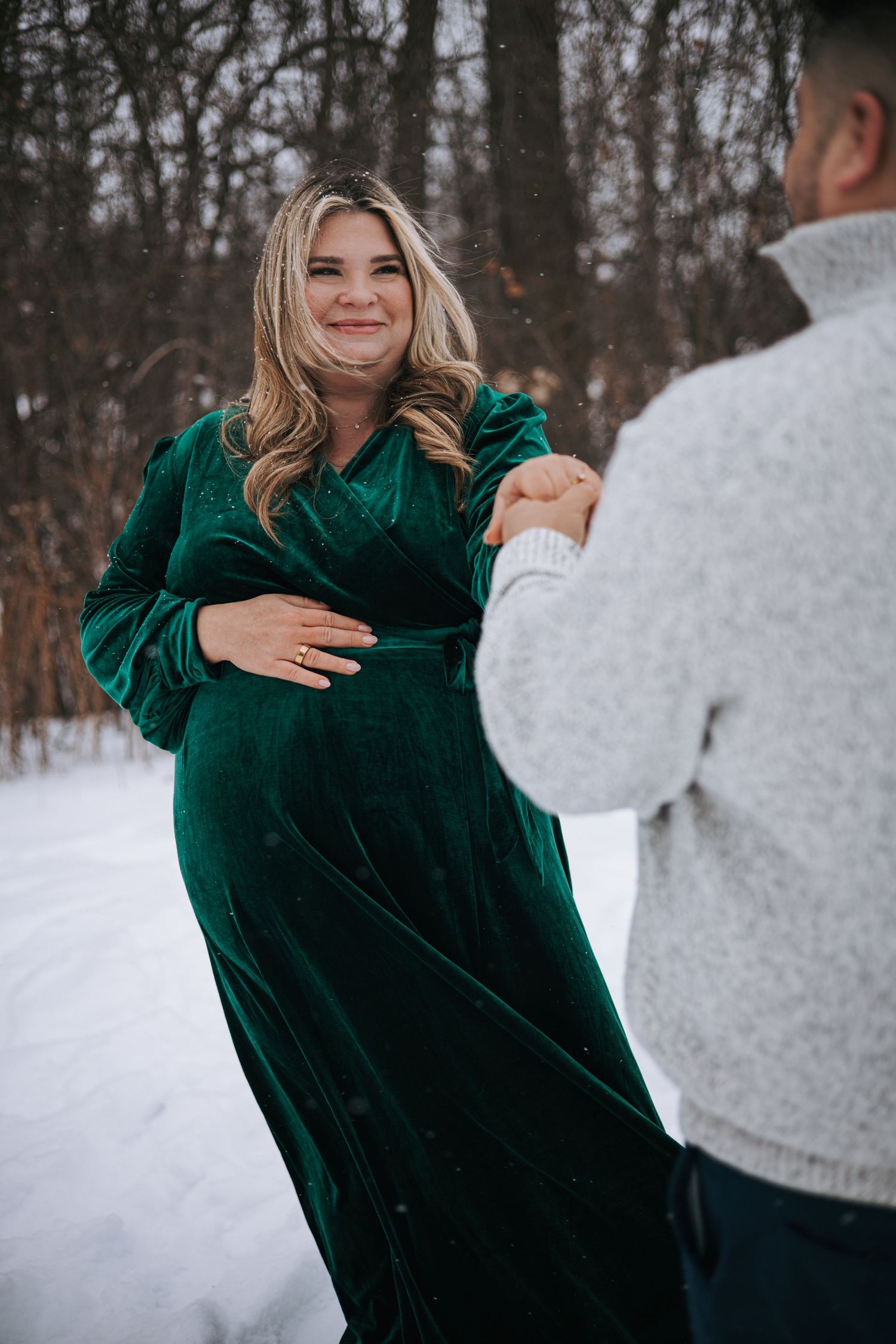 Pregnant woman in green velvet dress holding partners hand while smiling during snowy winter maternity photoshoot.