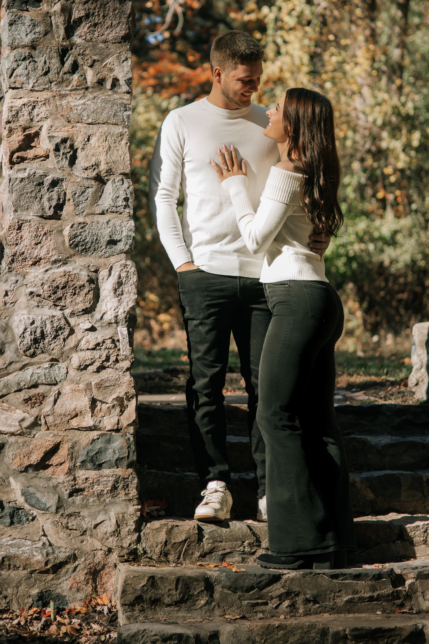 Engaged couple in matching white sweaters standing close on stone steps in autumn park.