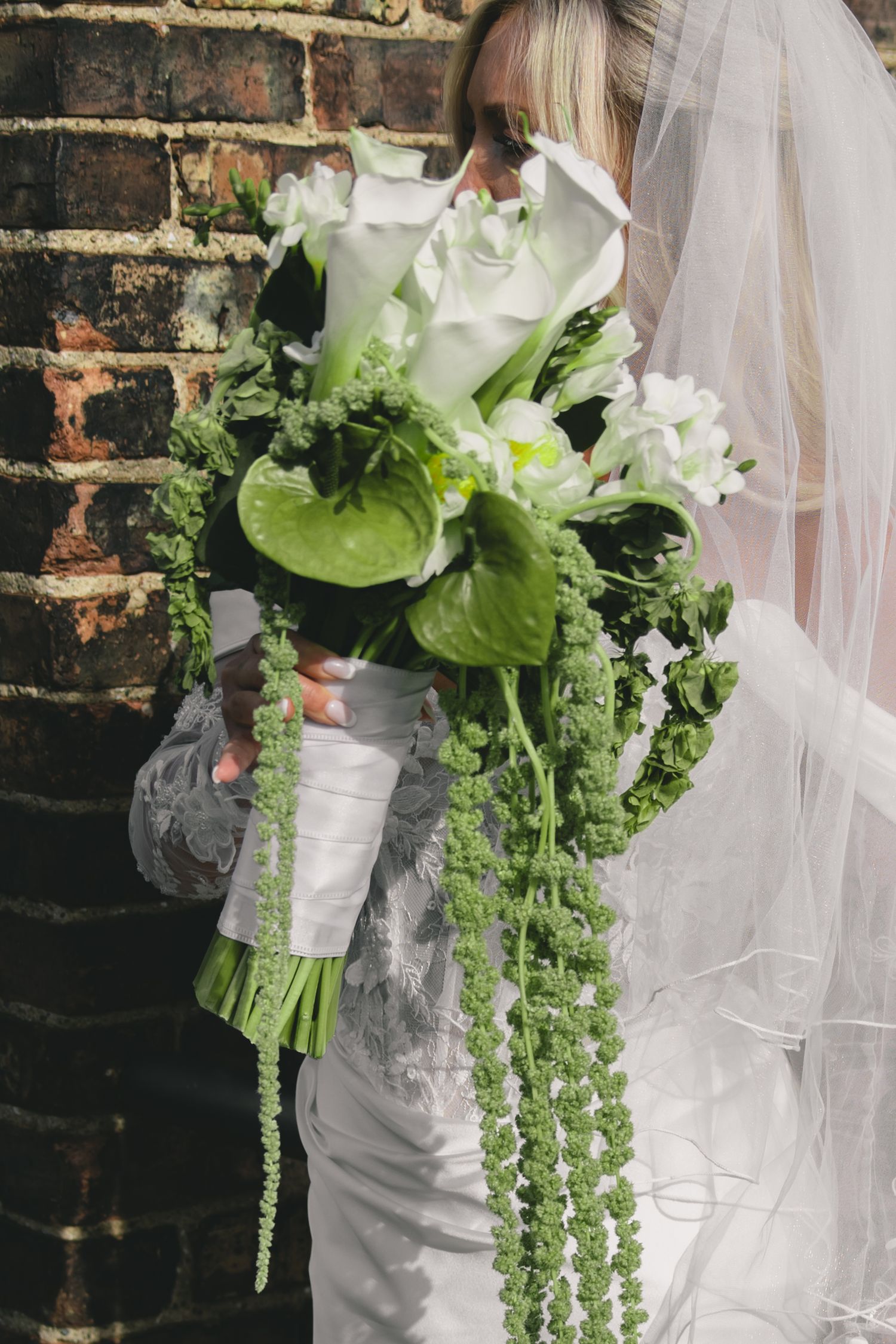 Bride holding a lush green and white wedding bouquet with cascading foliage against a brick wall.