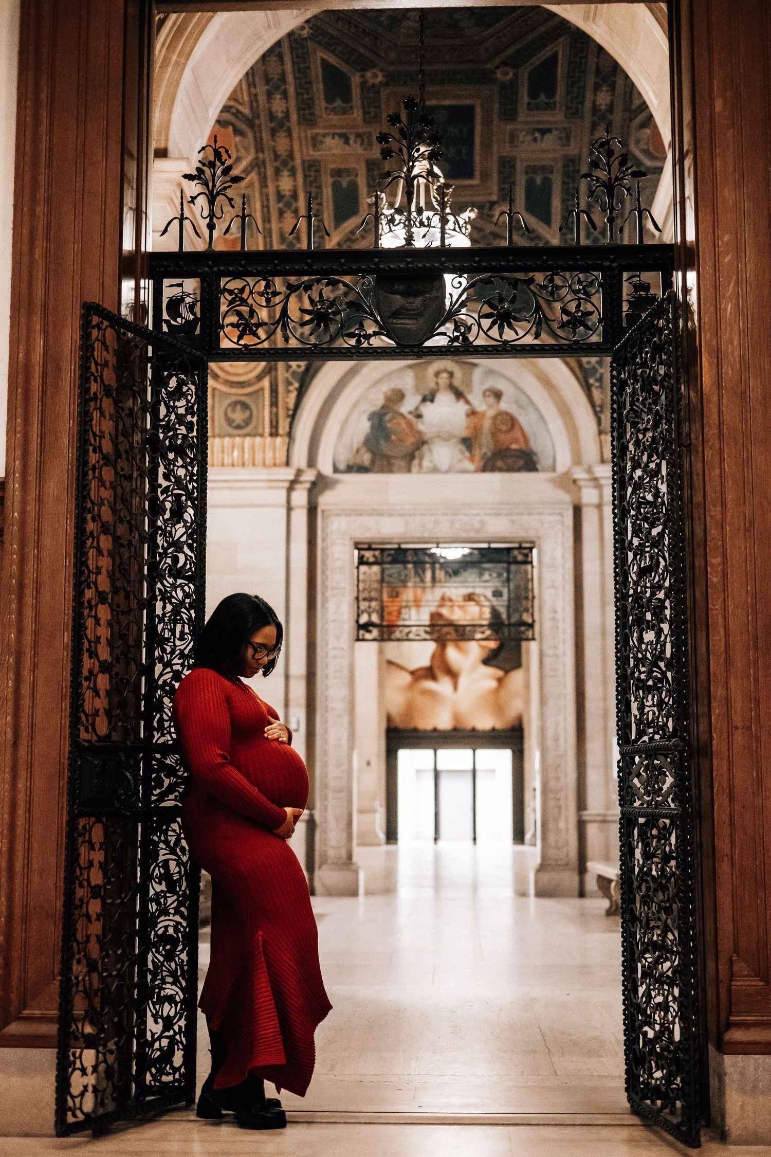 A pregnant woman in a red dress stands between ornate iron gates in a grand architectural hall.