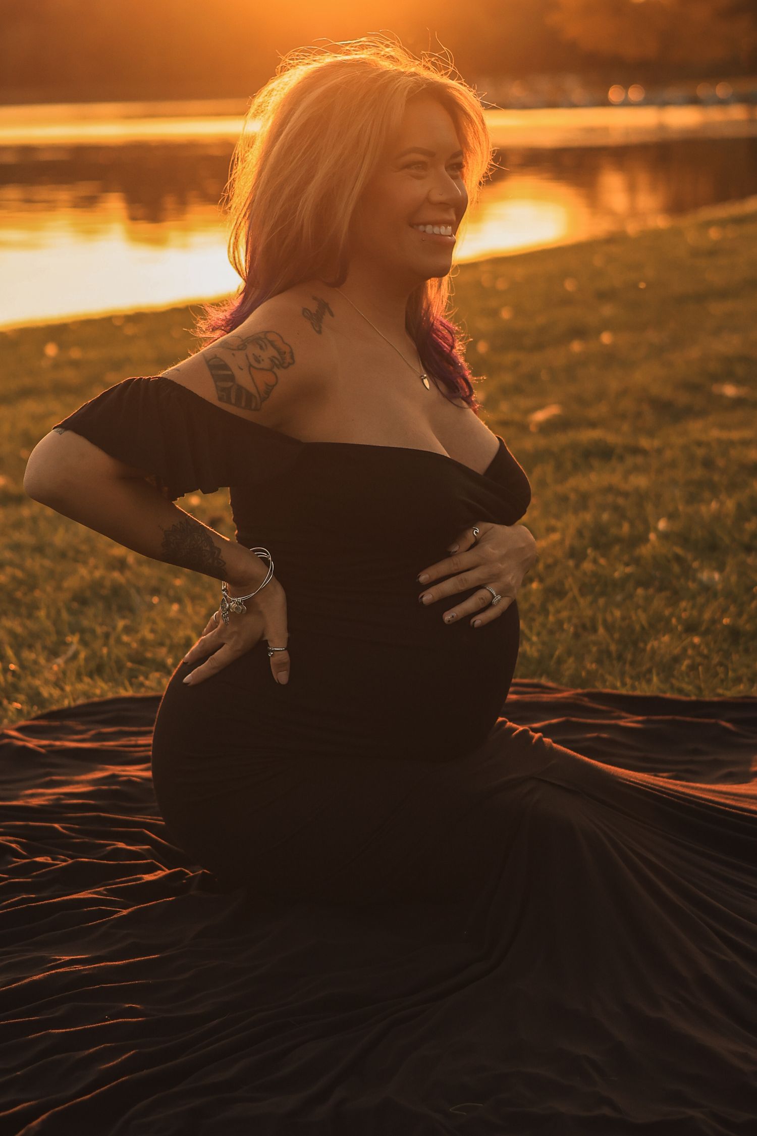 Radiant pregnant woman in off-shoulder black gown sitting outdoors smiling broadly in warm golden sunset light.