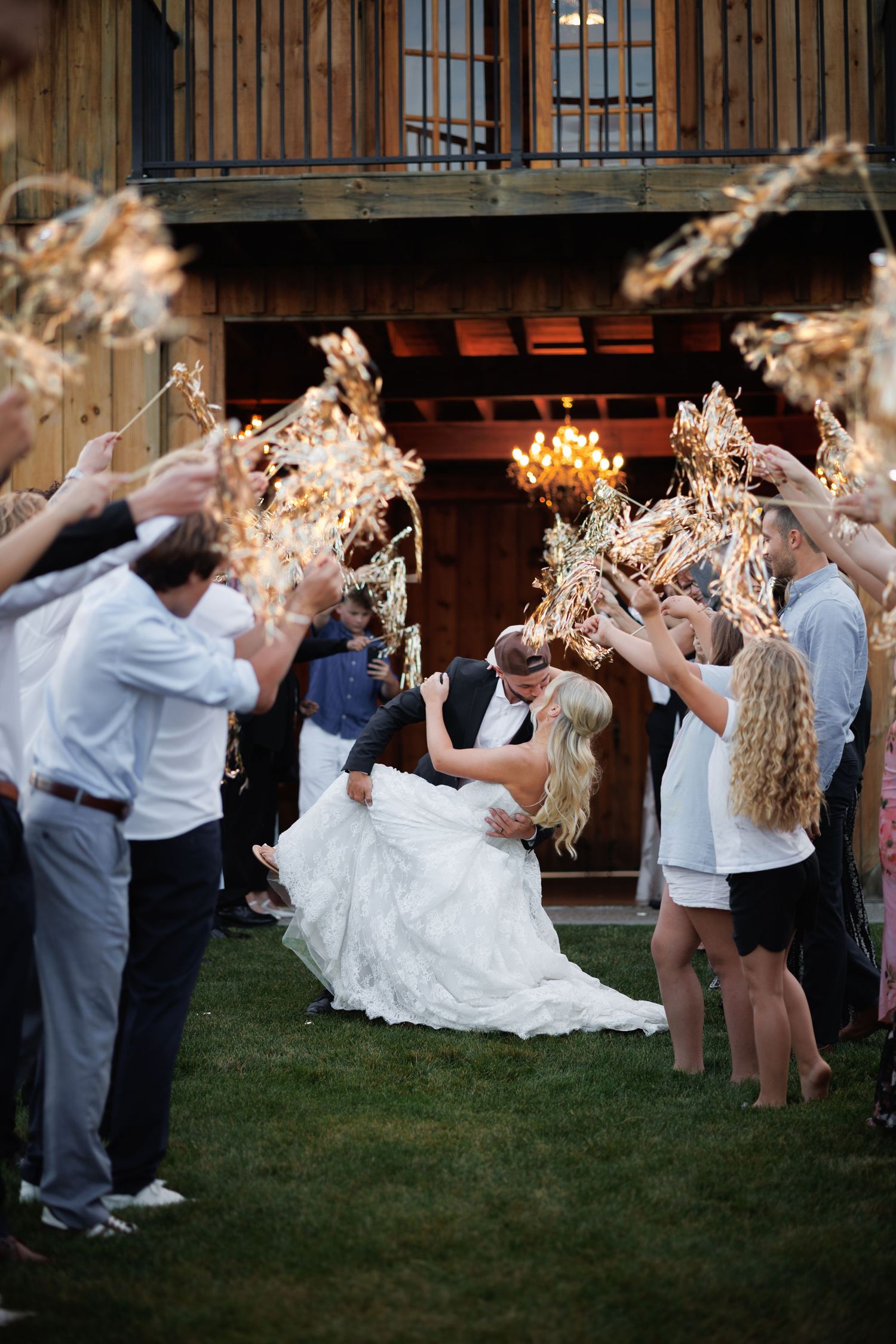 Bride and groom share a dip kiss during outdoor wedding exit while guests hold dried floral arrangements aloft.