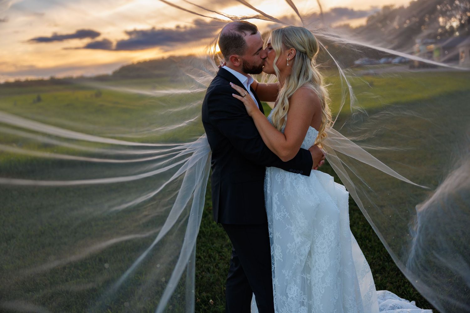 Bride and groom sharing a romantic kiss under flowing veil at sunset on a lush green hillside.