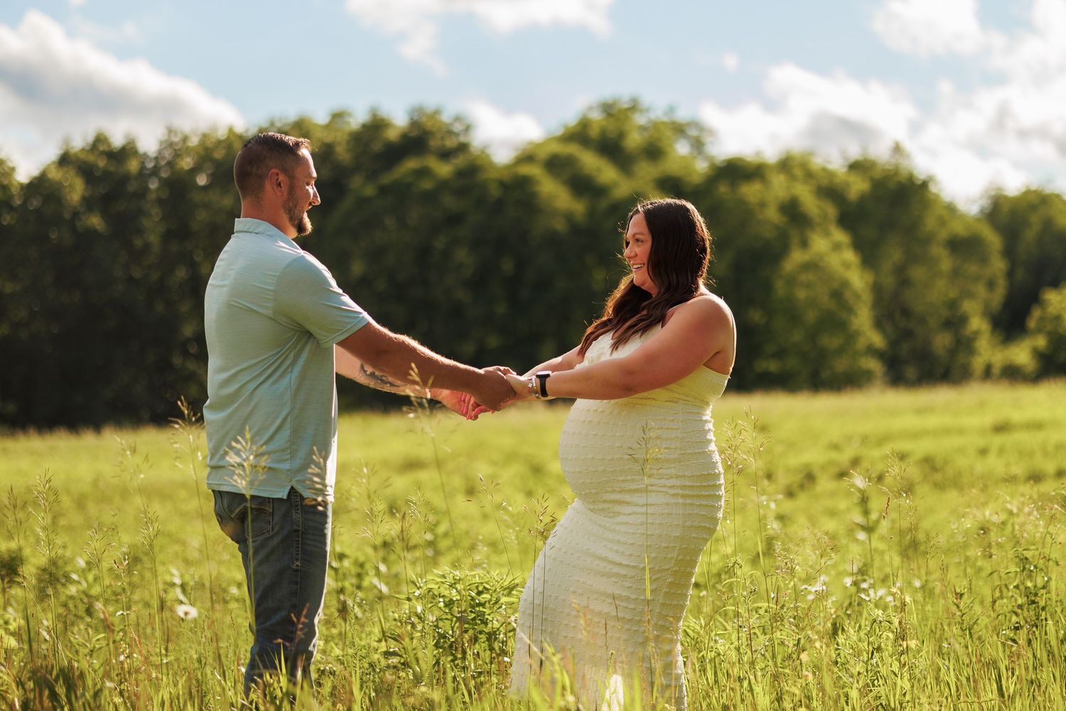 Pregnant couple holding hands in a sunny green field, sharing a tender moment outdoors with trees in the background.