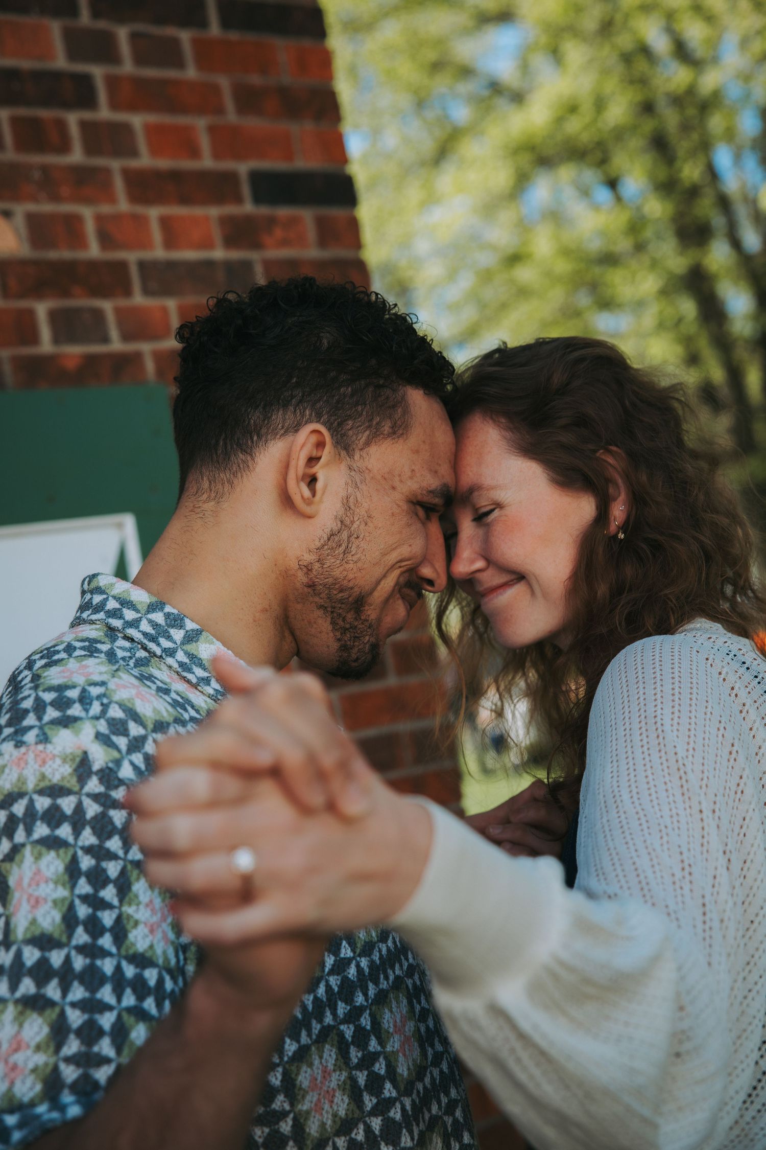 Engaged couple dancing closely forehead to forehead, holding hands near a brick wall with trees in the background.