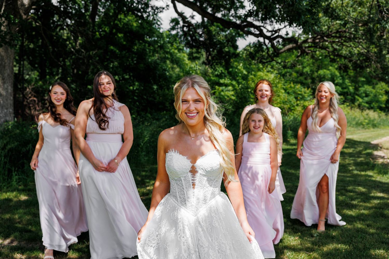Smiling bride in strapless gown poses with bridesmaids in blush dresses outdoors.