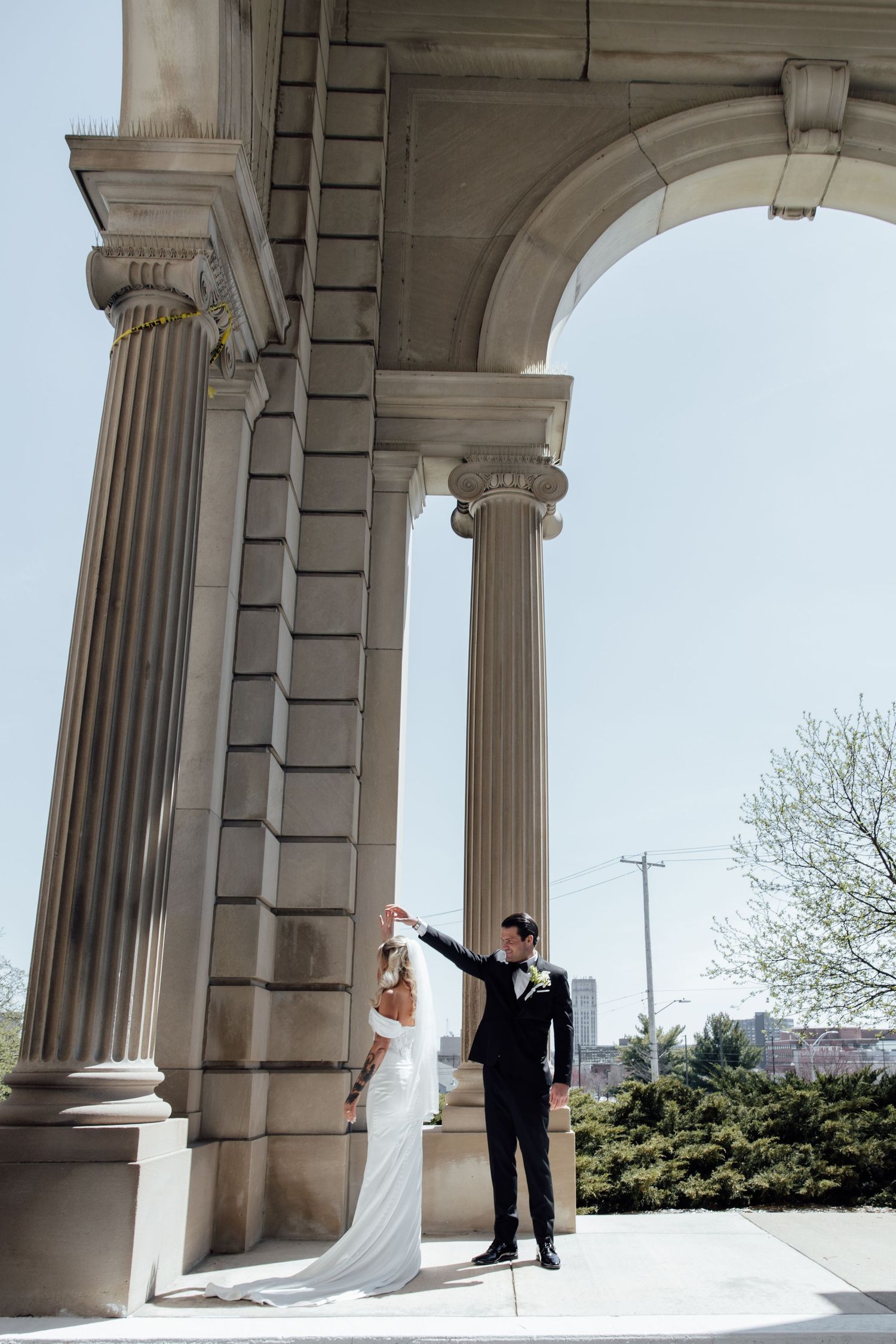Groom twirling bride in elegant white wedding dress beneath ornate stone arches and tall classical columns.