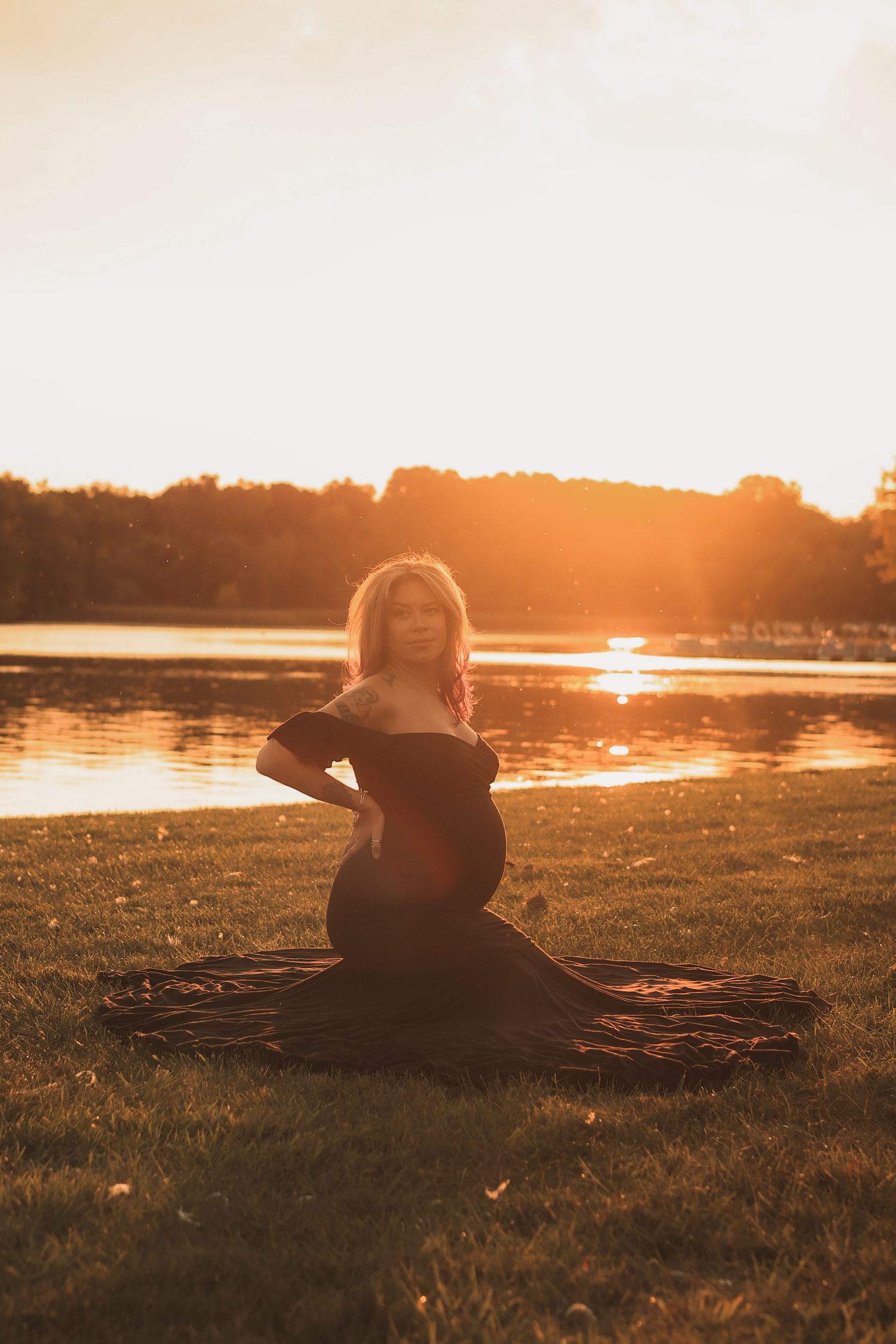 Pregnant woman in flowing black maternity gown sitting on grass by lake at sunset.