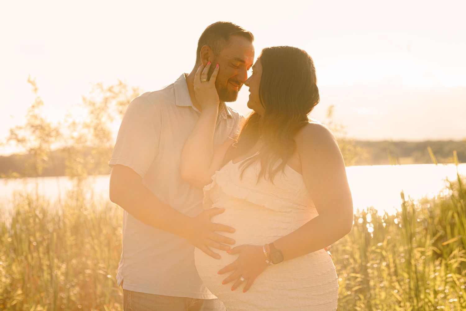 Couple sharing a tender moment at golden hour, man touching woman's pregnant belly near a sunlit lake.