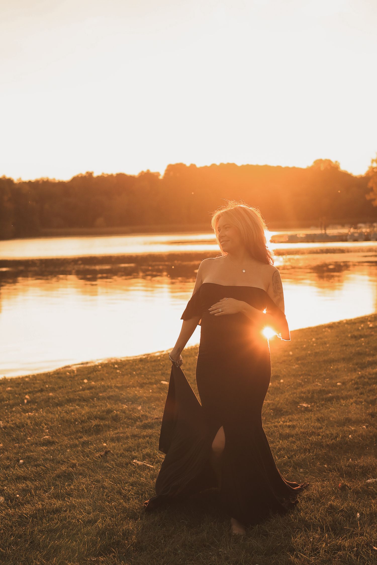 Pregnant woman in black off-shoulder gown walking by lake holding dress train at sunset.