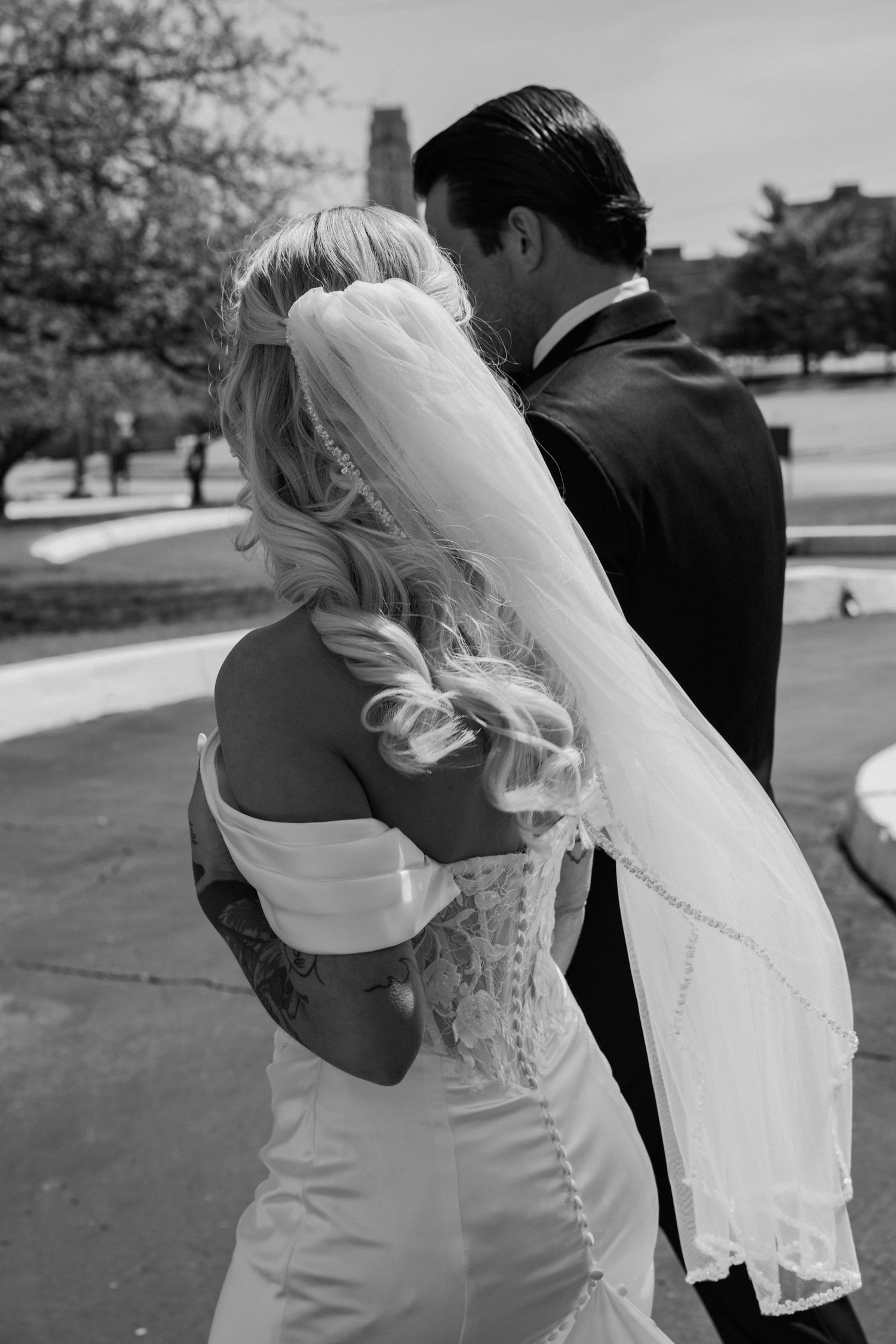 Black and white photo of bride with lace corset gown and flowing veil walking with groom outdoors.