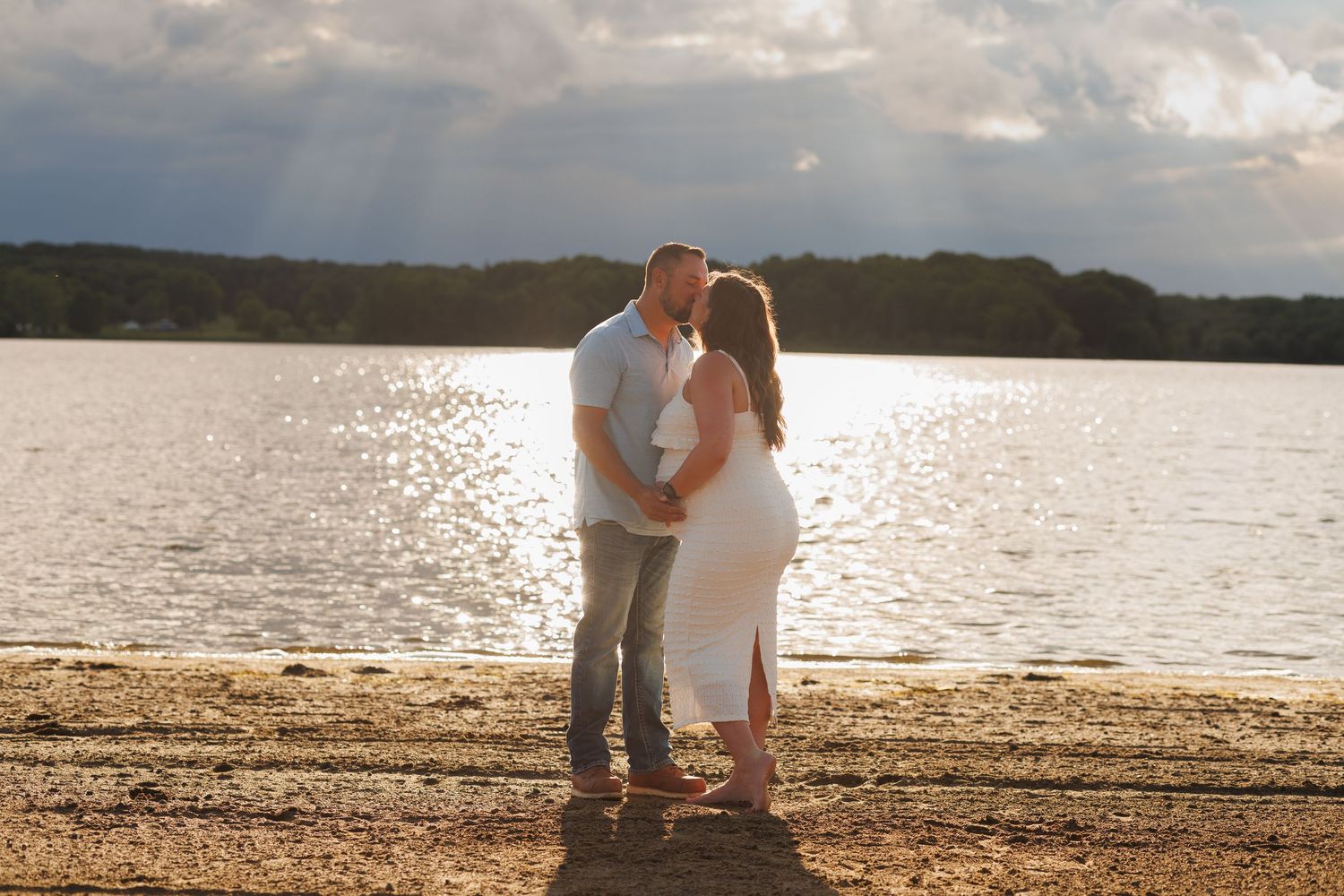 Couple sharing a romantic kiss on a sandy beach at golden hour with sunrays over a calm lake.
