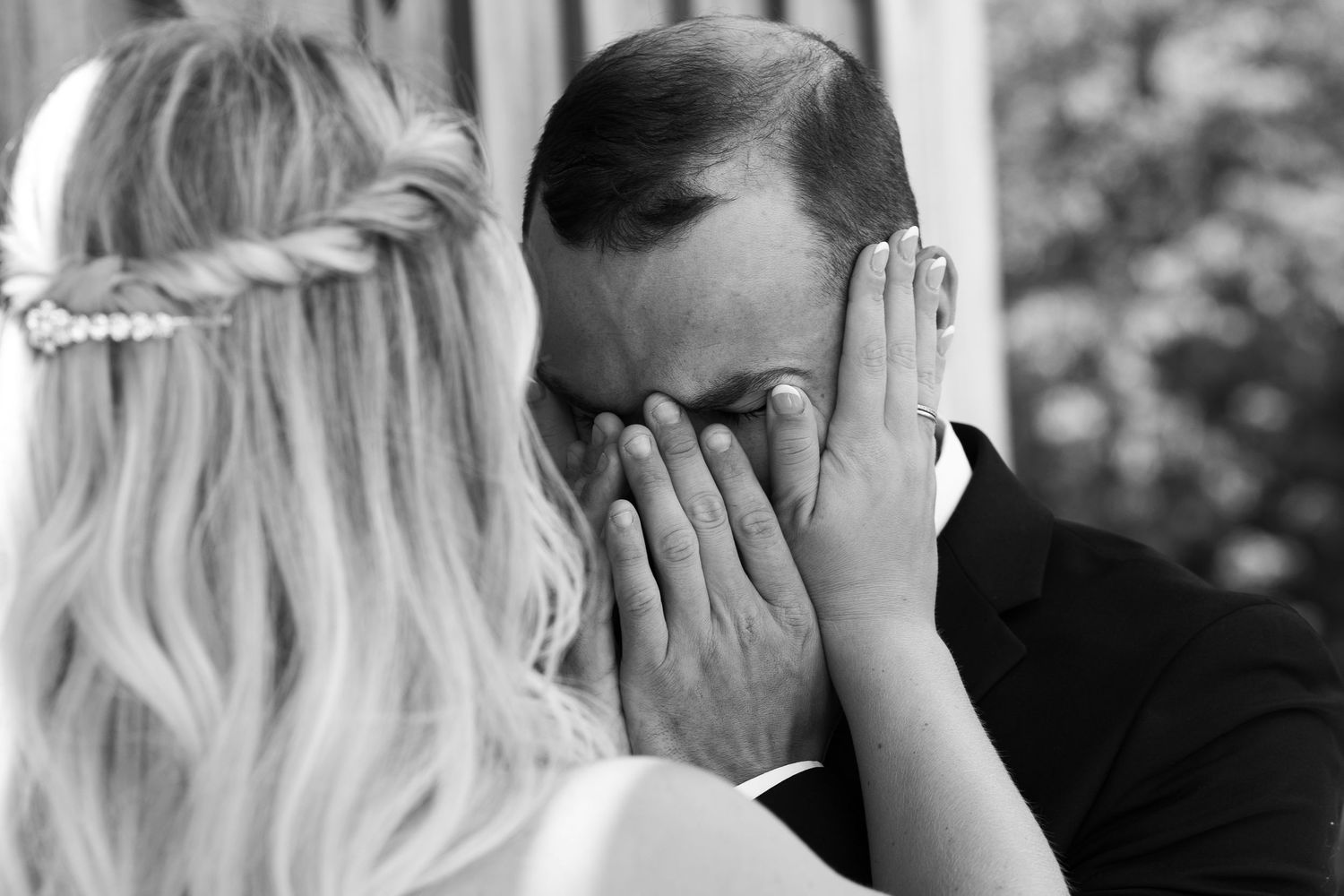Black and white photo of emotional groom covering his face with hands as bride approaches during outdoor wedding ceremony.