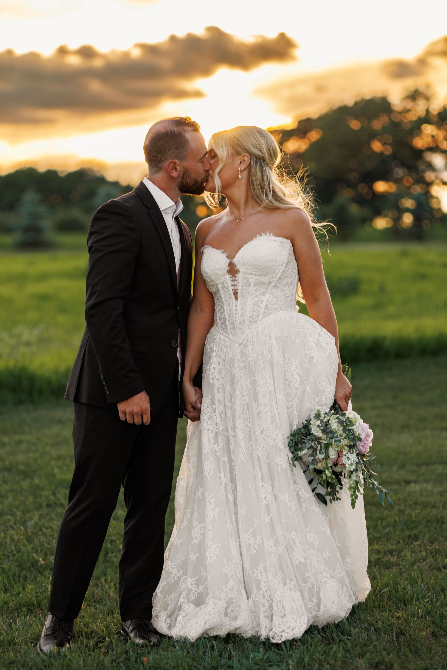 Bride and groom share a romantic kiss at sunset in a lush green field, bride holding a bouquet in lace gown.