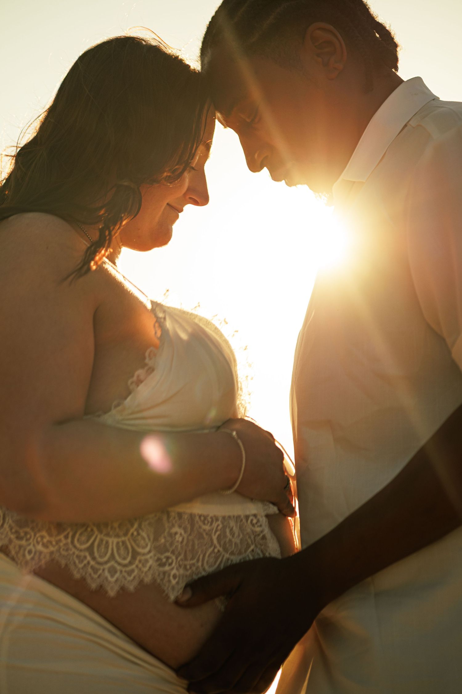 Pregnant couple sharing intimate moment backlit by golden sunset light, woman in white lace dress with partner touching baby bump.
