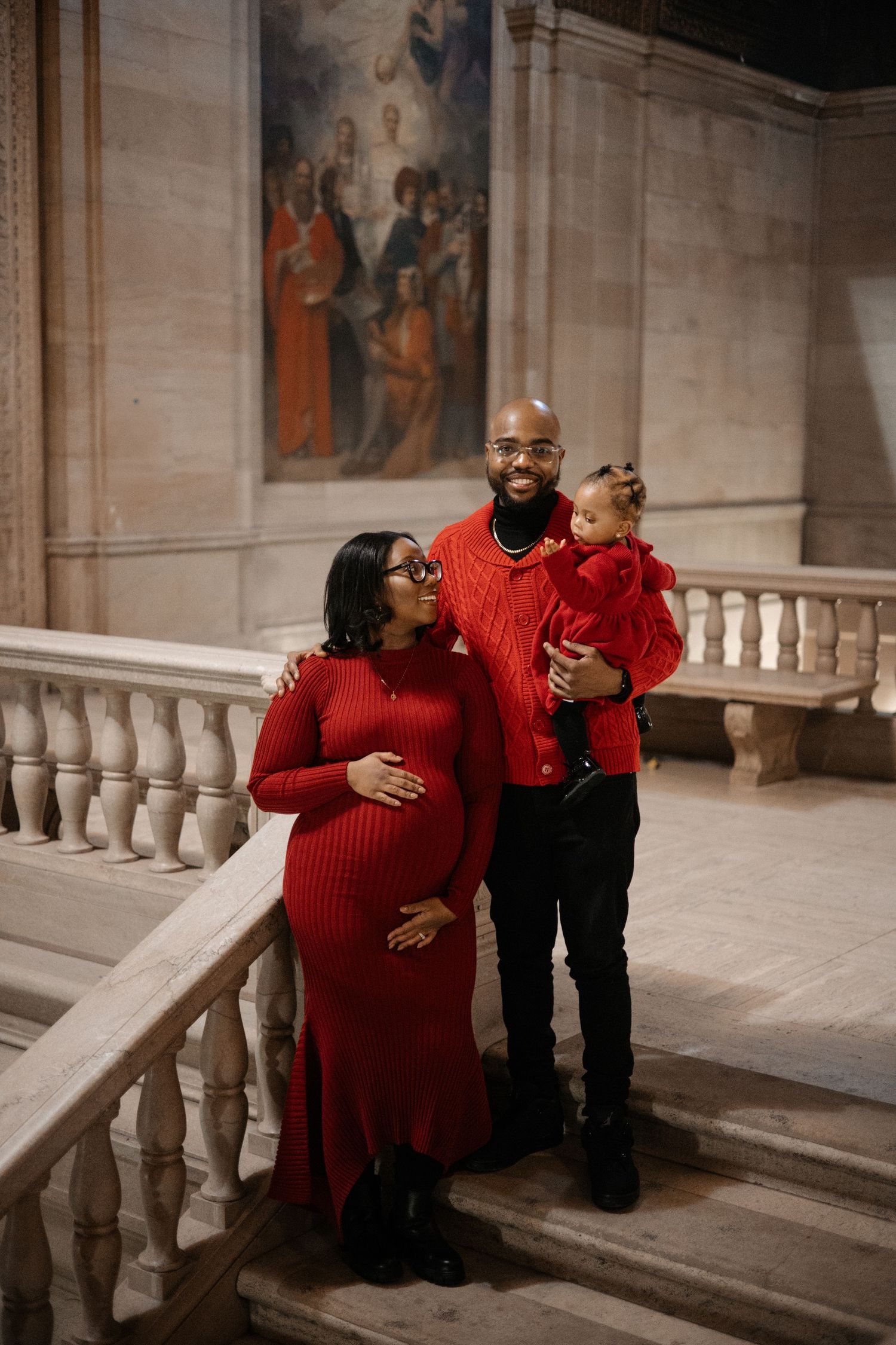 A pregnant woman in red poses with her partner and young child on marble stairs in a grand hall.