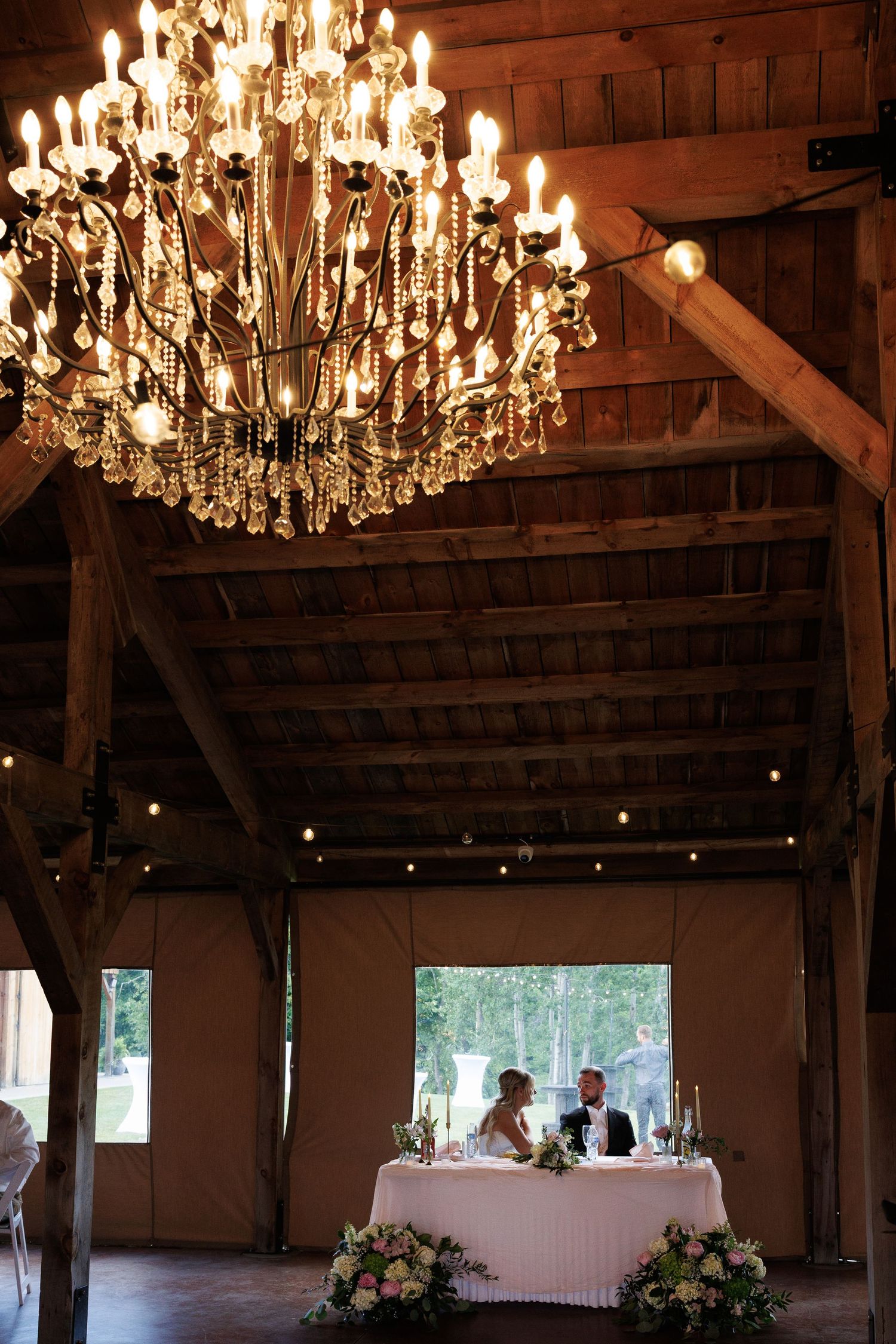 Elegant barn wedding reception with crystal chandelier illuminating couple at sweetheart table with floral arrangements.