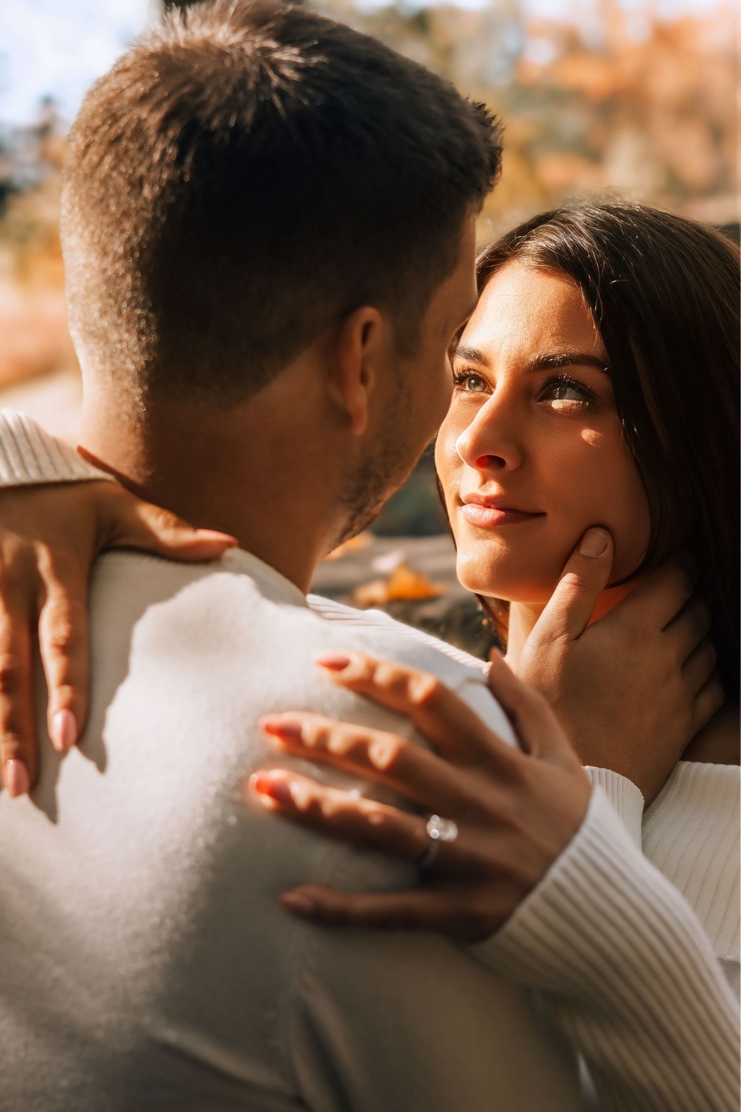 Romantic couple gazing into each others eyes outdoors, woman displaying diamond engagement ring in warm golden light.