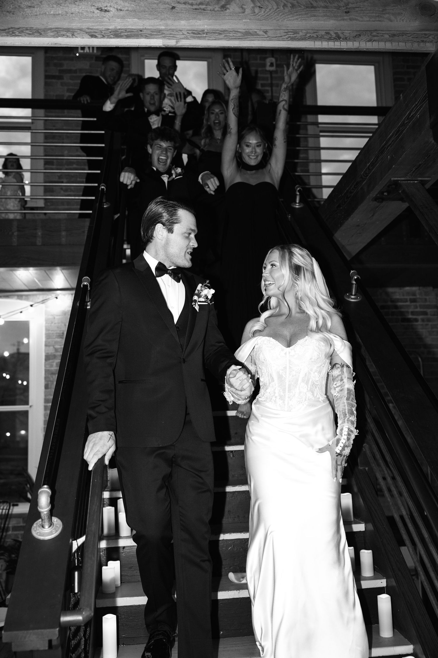 Bride and groom descend staircase hand in hand while wedding party celebrates behind them in black and white photo.