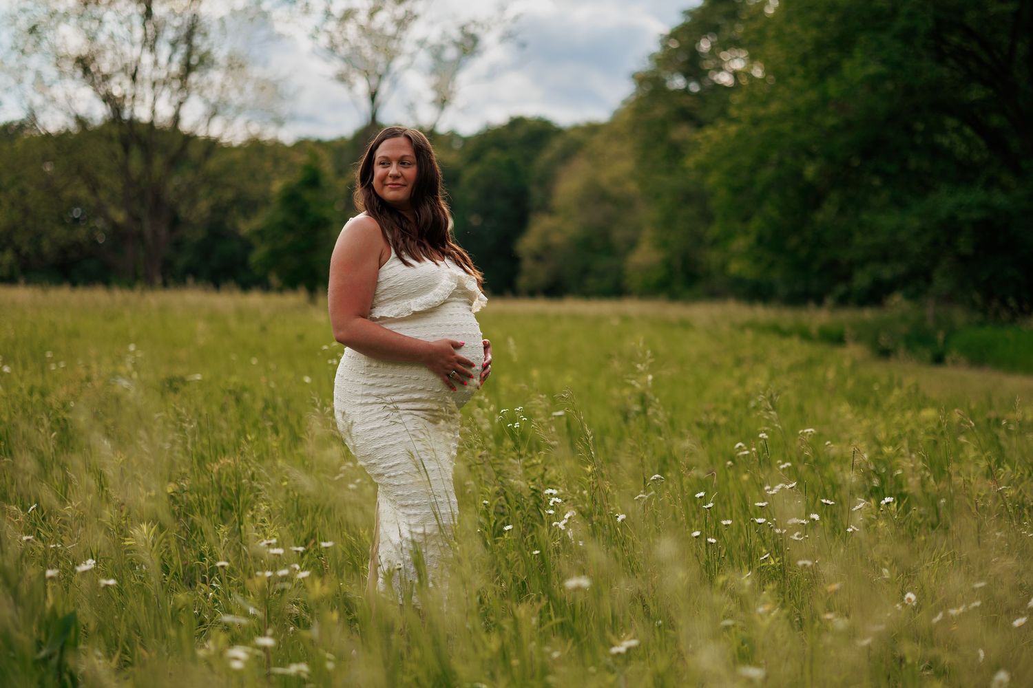 Pregnant woman in white dress cradling baby bump in a lush green meadow with wildflowers and trees.