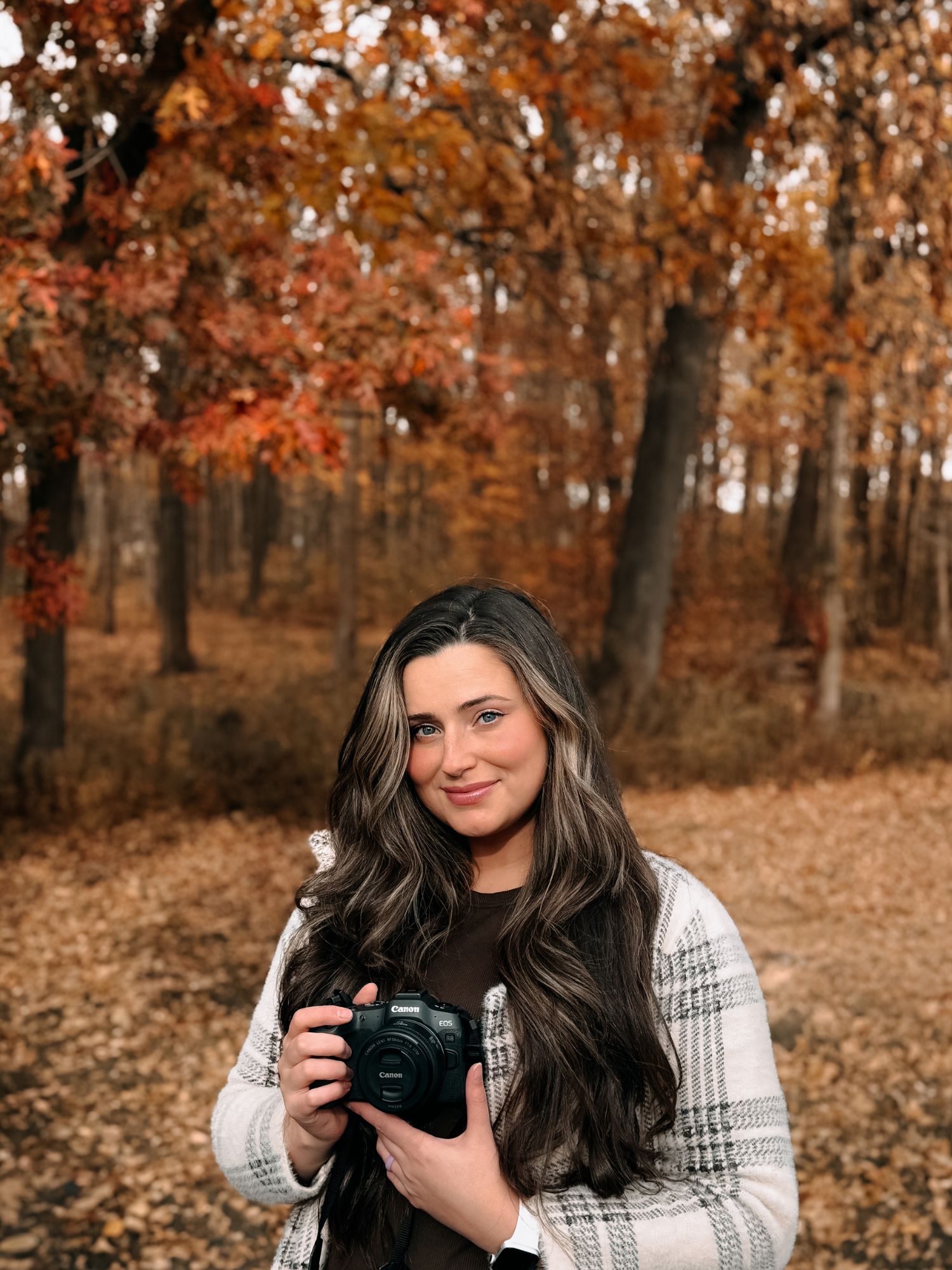 Woman with dark hair holding a camera in an autumn forest with orange and red foliage.