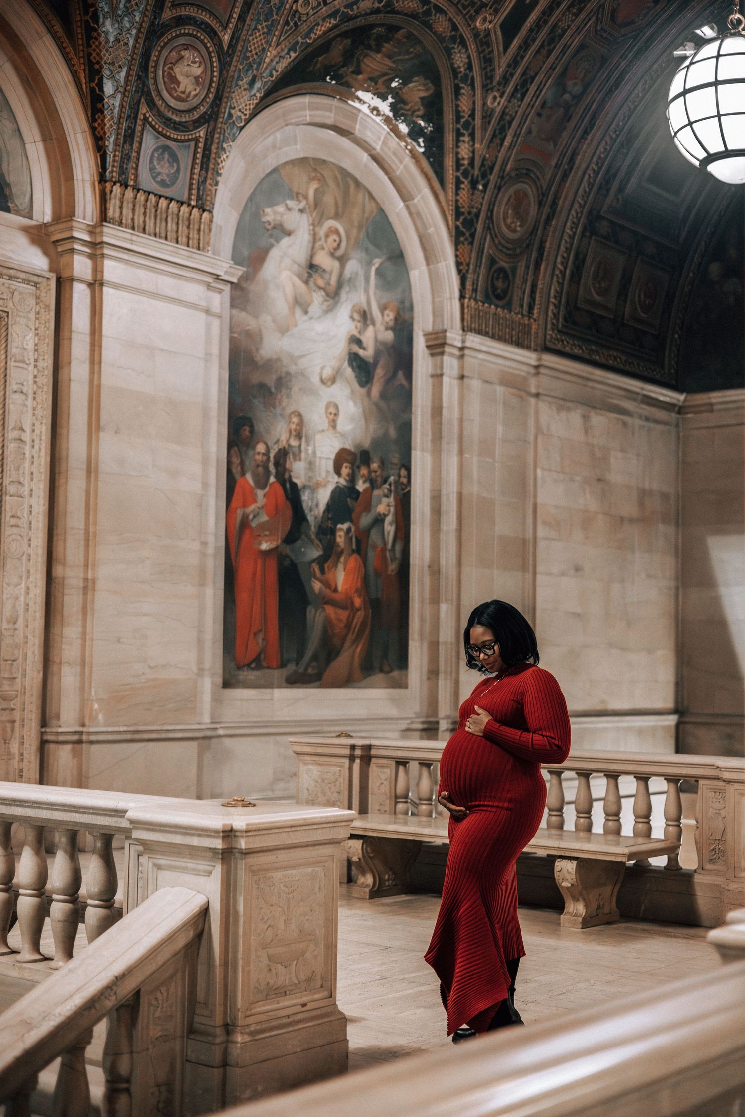 Elegant pregnant woman in red dress walking near marble balustrade in grand historic building.