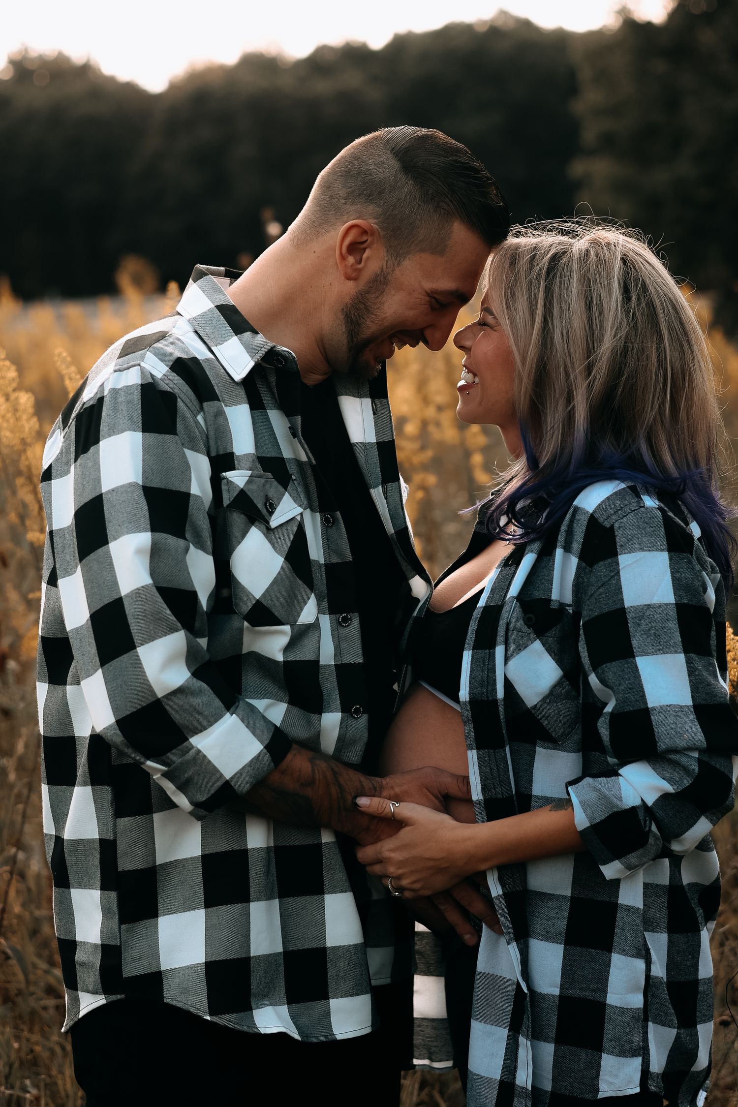 Pregnant couple in matching plaid shirts embracing in a golden field during a maternity photo session.