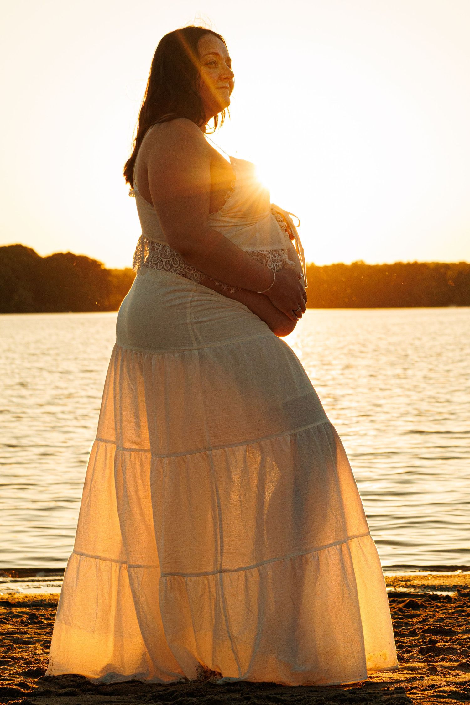 Pregnant woman in white lace gown cradling belly at golden sunset by lake shore.