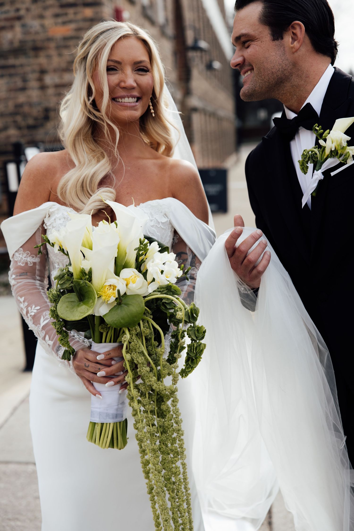 Smiling blonde bride in off-shoulder lace gown holds cascading green floral bouquet with groom.