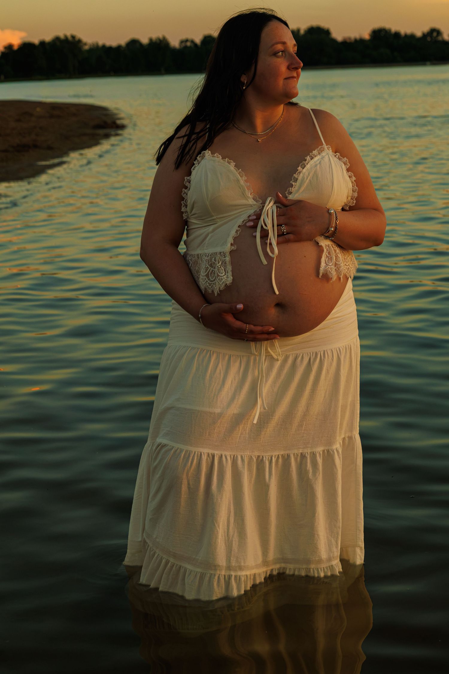 Pregnant woman in white dress standing in lake water at sunset, cradling her baby bump.