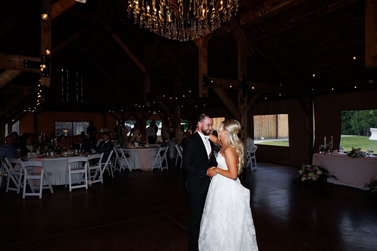 Bride and groom share first dance in rustic barn wedding venue with chandelier and string lights.