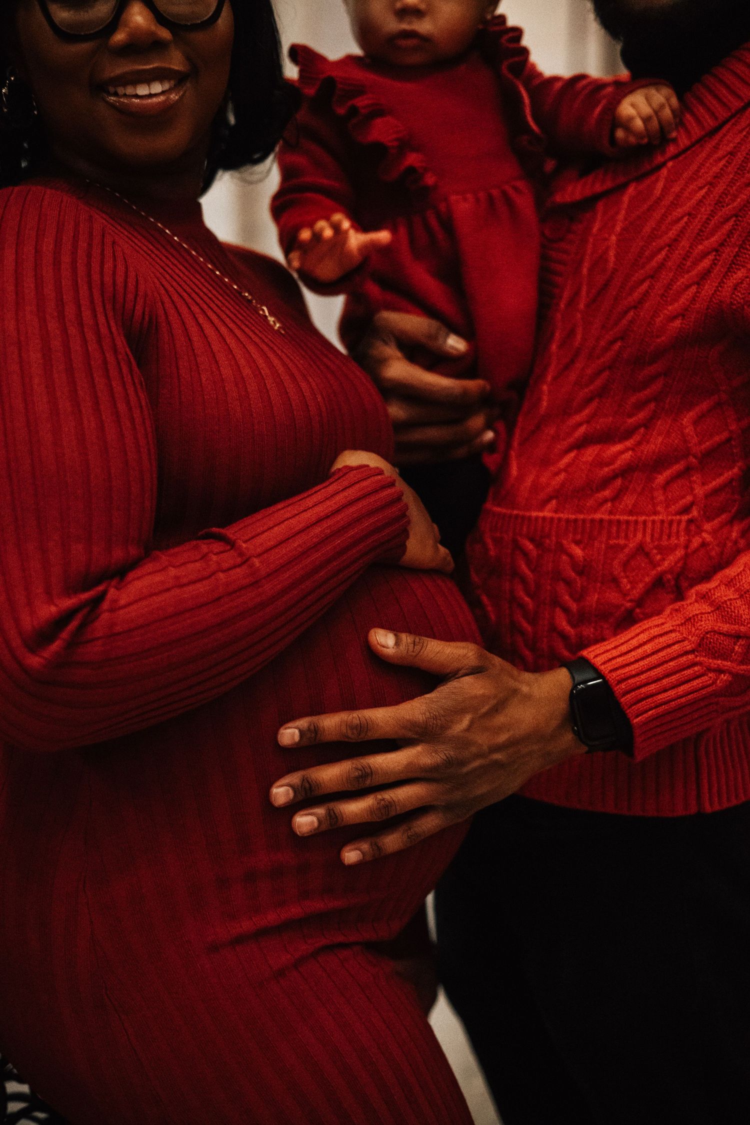 Family in coordinating red outfits with father holding toddler and hand on pregnant mother's belly.
