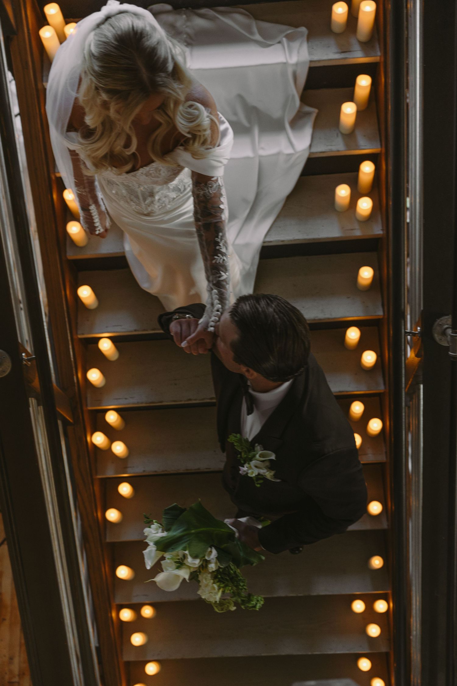Aerial view of bride and groom on candlelit staircase, groom kissing bride's hand romantically.