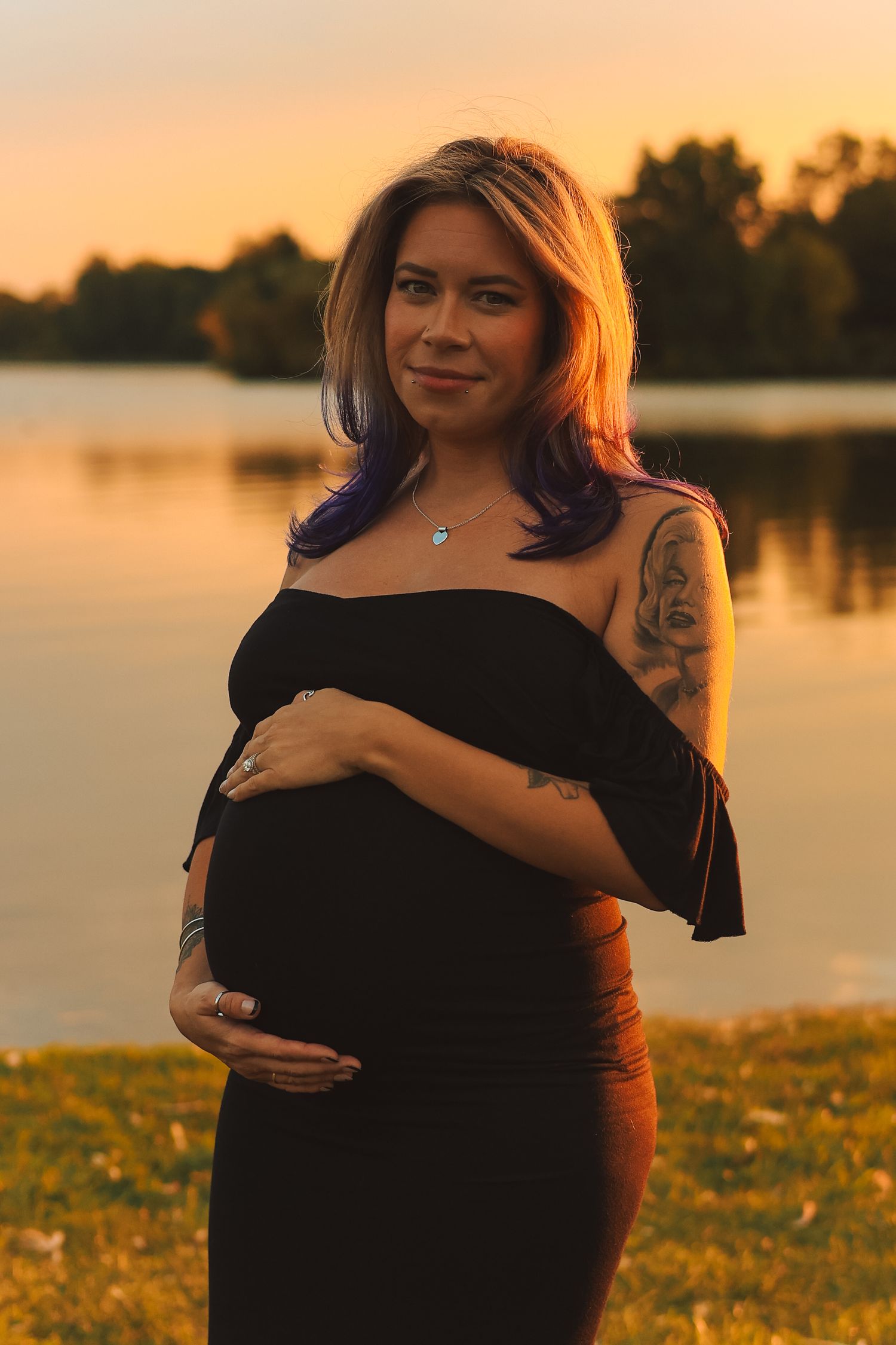 Tattooed pregnant woman in black dress smiling while cradling belly at golden sunset lakeside.