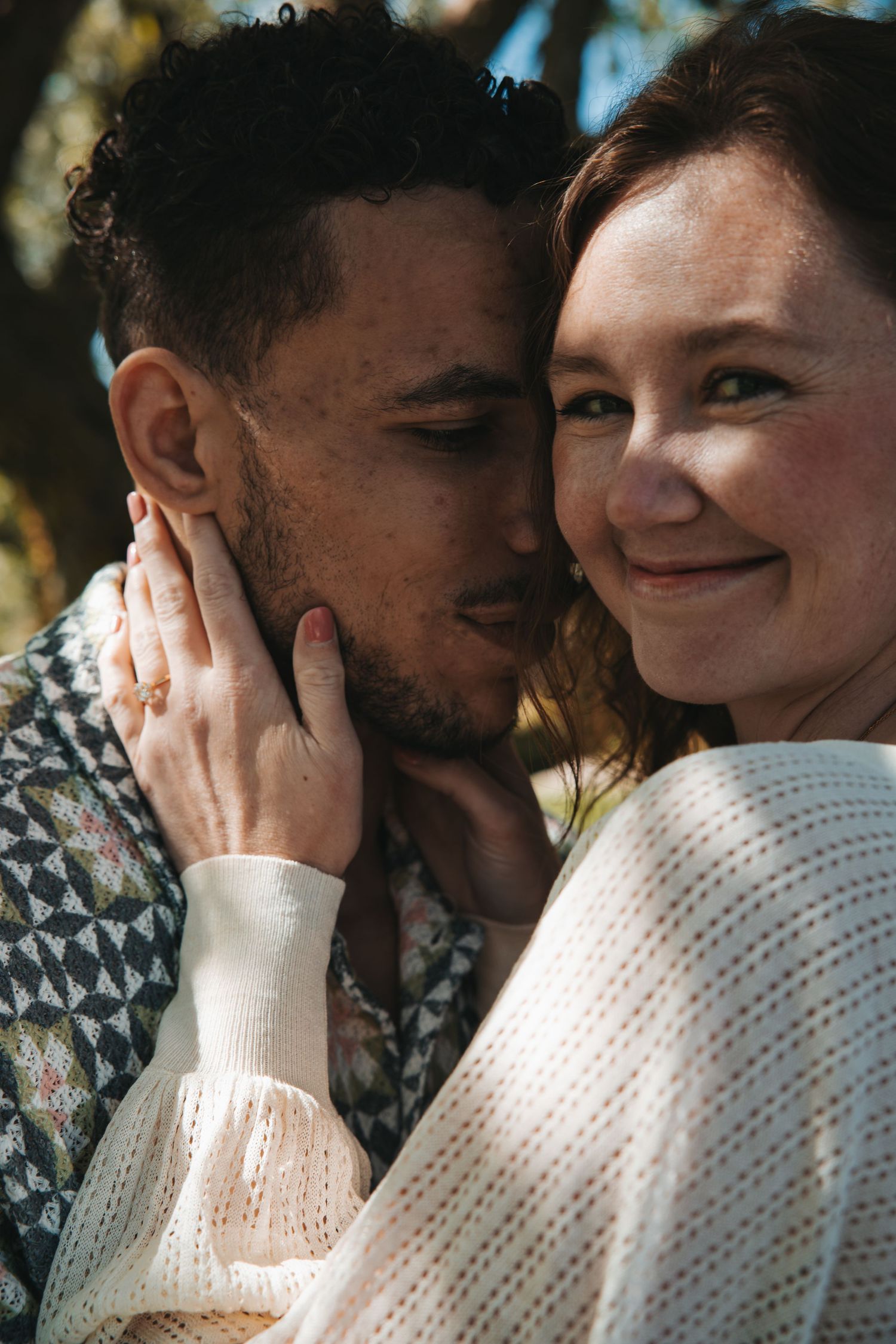 Woman smiling at camera while man kisses her cheek, she gently holds his face showing off her engagement ring.