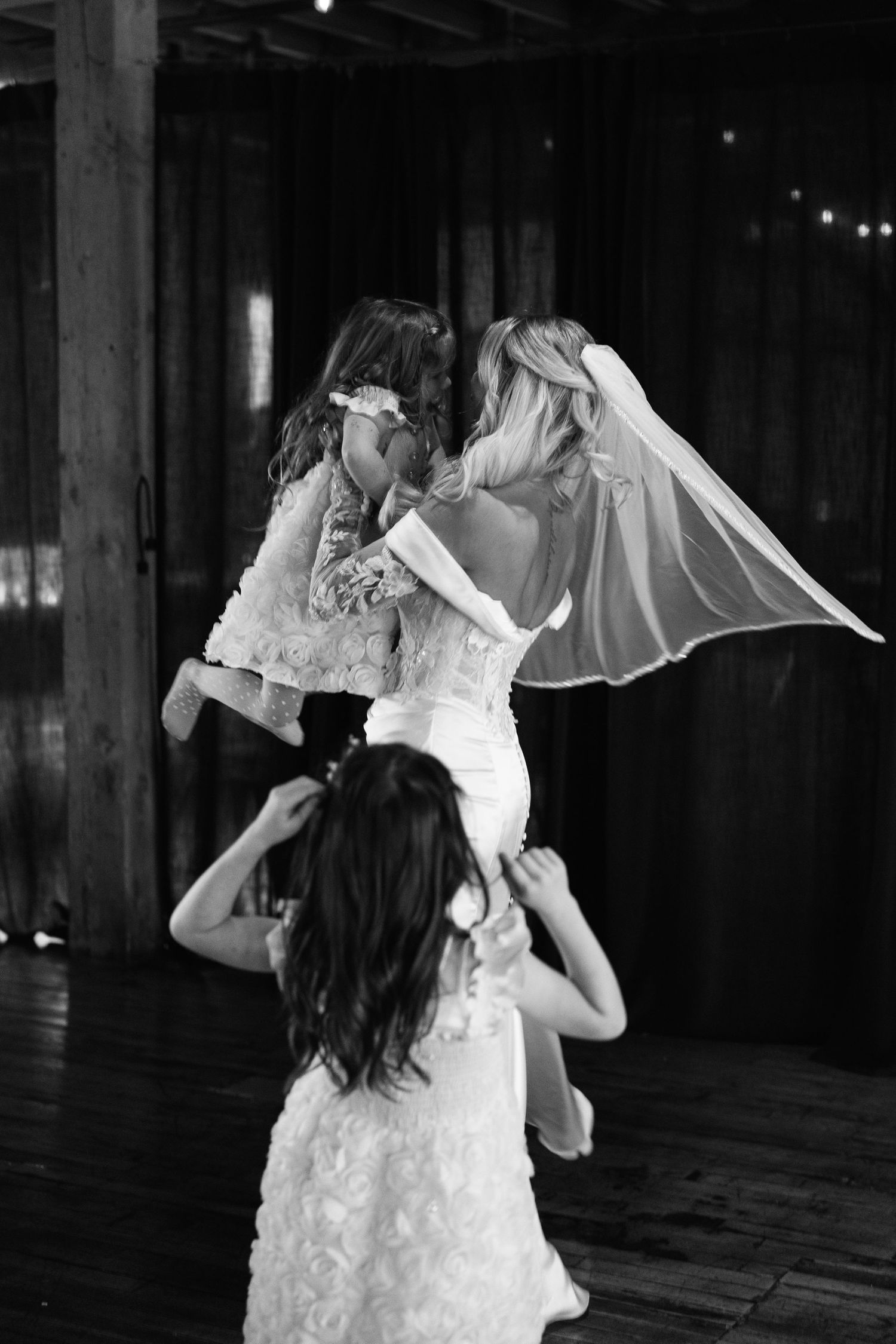 Bride lifts a flower girl while another child watches in a black and white wedding photo.