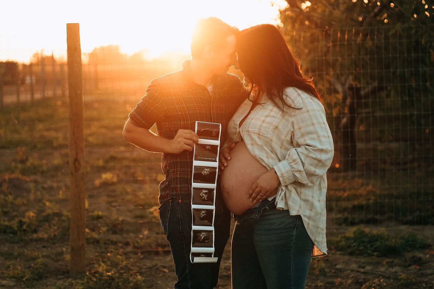 A couple kisses while holding ultrasound photos during a golden hour outdoor maternity photoshoot.